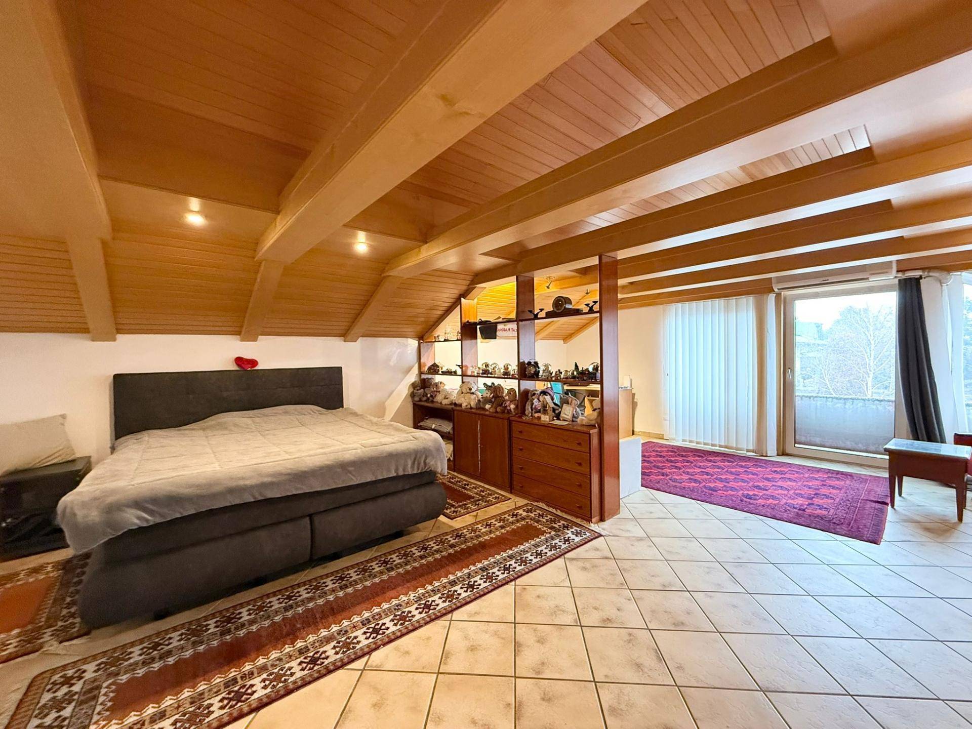 Geräumiges Schlafzimmer im Dachgeschoss mit Holzbalkendecke, Fliesenboden und Zugang zum Balkon.