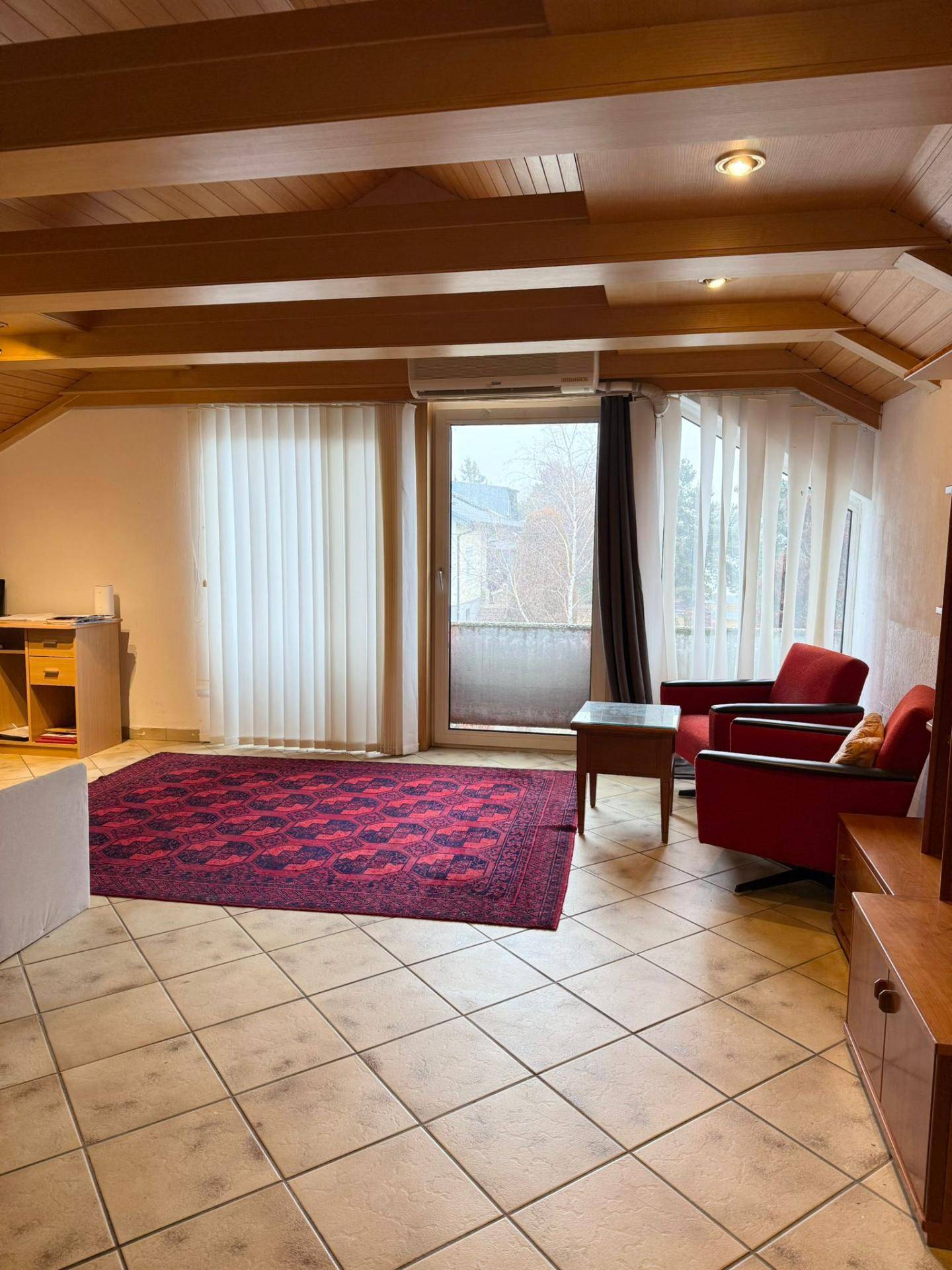 Geräumiger Wohnbereich mit Fliesenboden, Holzbalkendecke und Zugang zu einem Balkon.