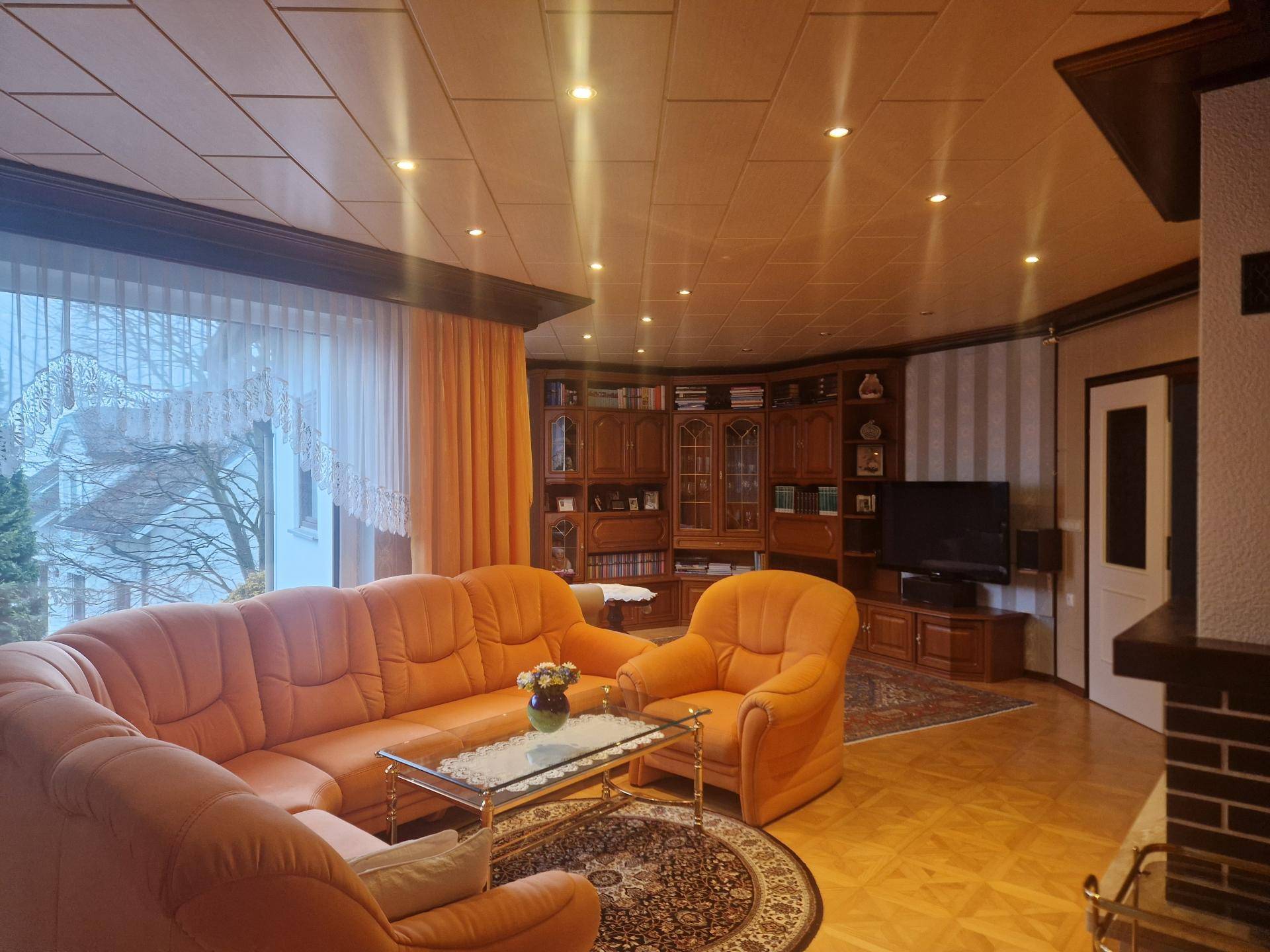 Wohnzimmer mit bequemen orangefarbenen Sofas, großem Holzschrank und Blick ins Grüne.