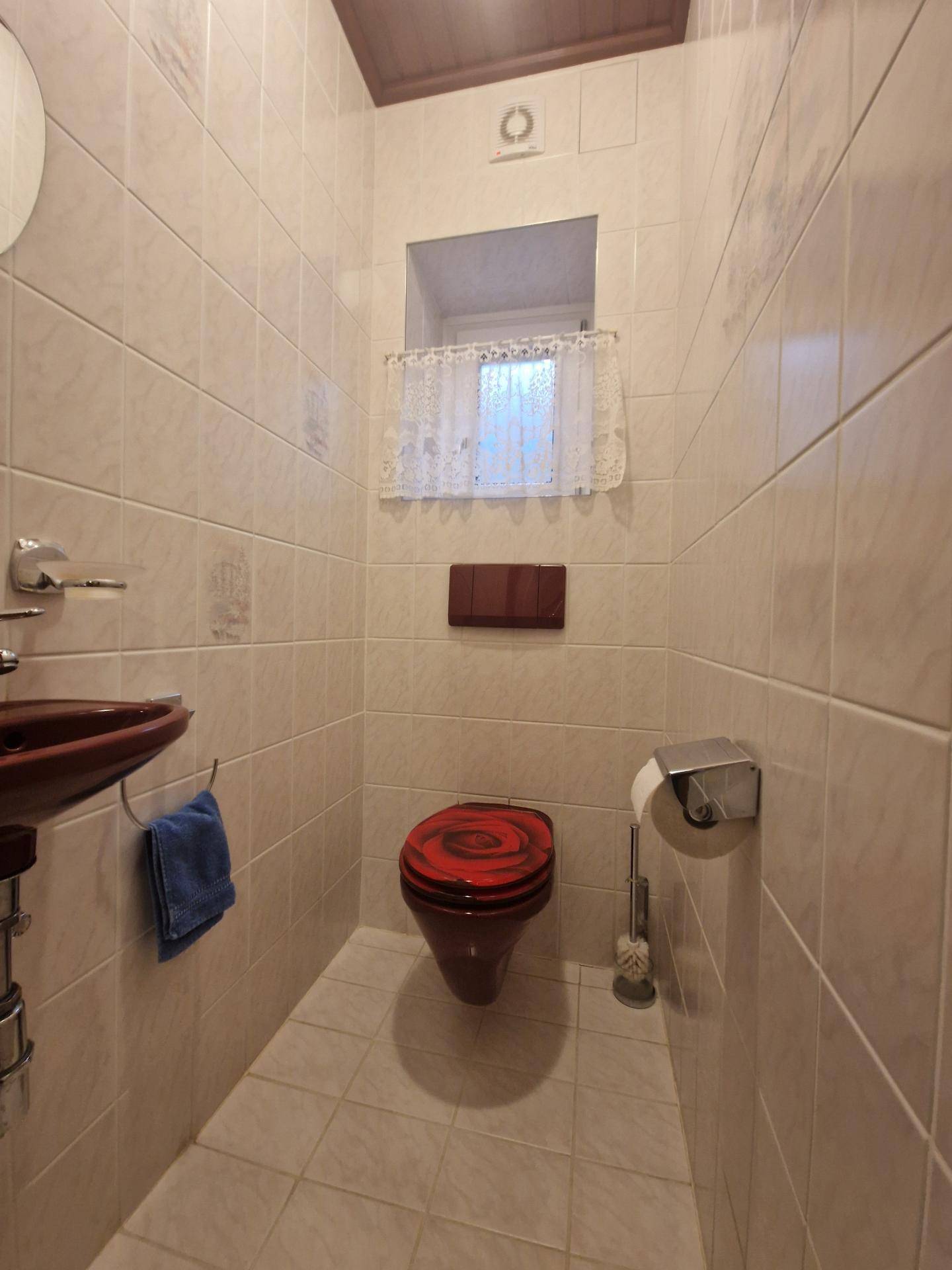 Kleines Gäste-WC mit Fliesen, rotem Toilettensitz und Fenster für natürliche Belüftung.