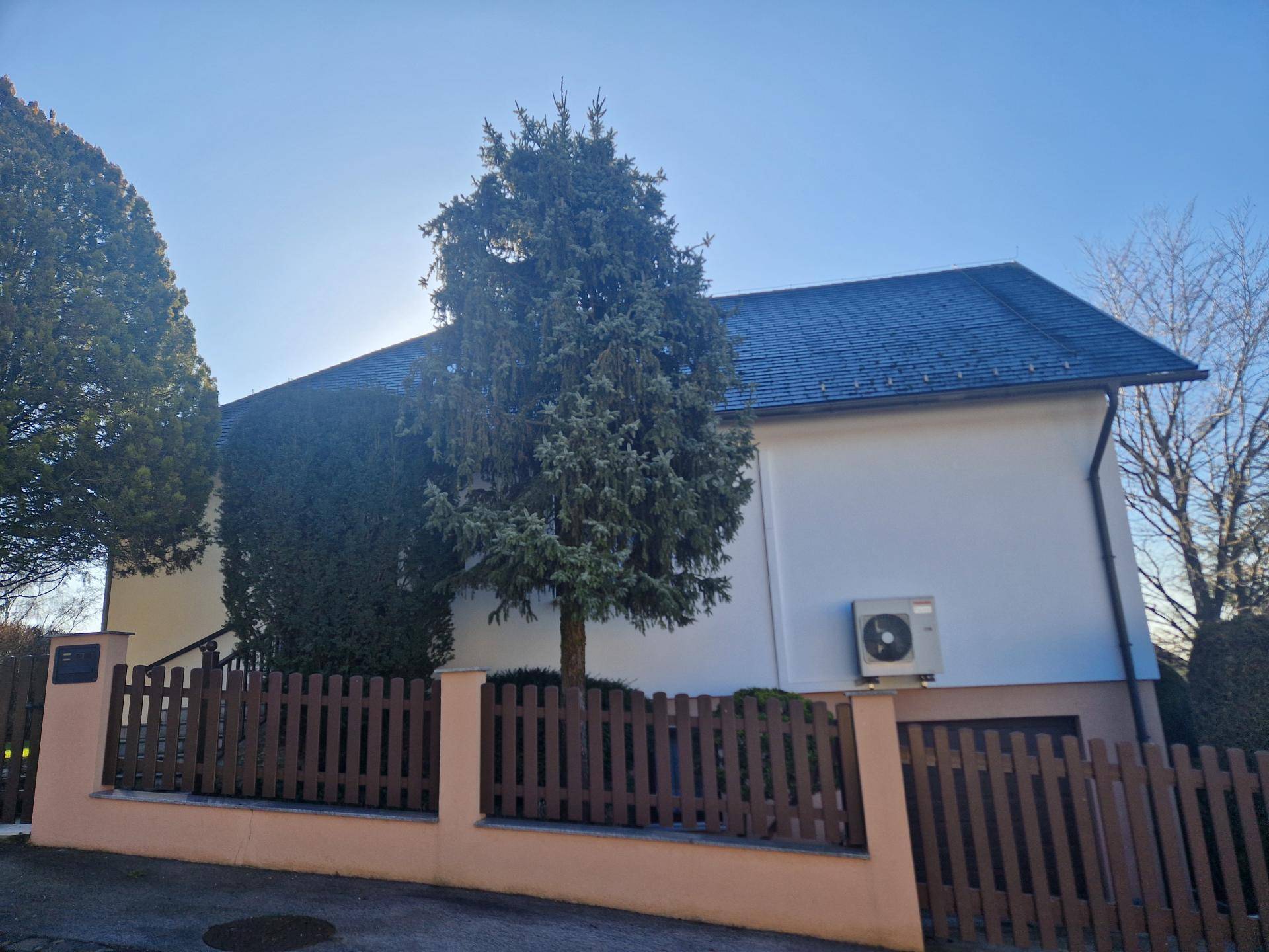Seitenansicht des Hauses mit Zaun, hohen Bäumen und einer Außeneinheit.