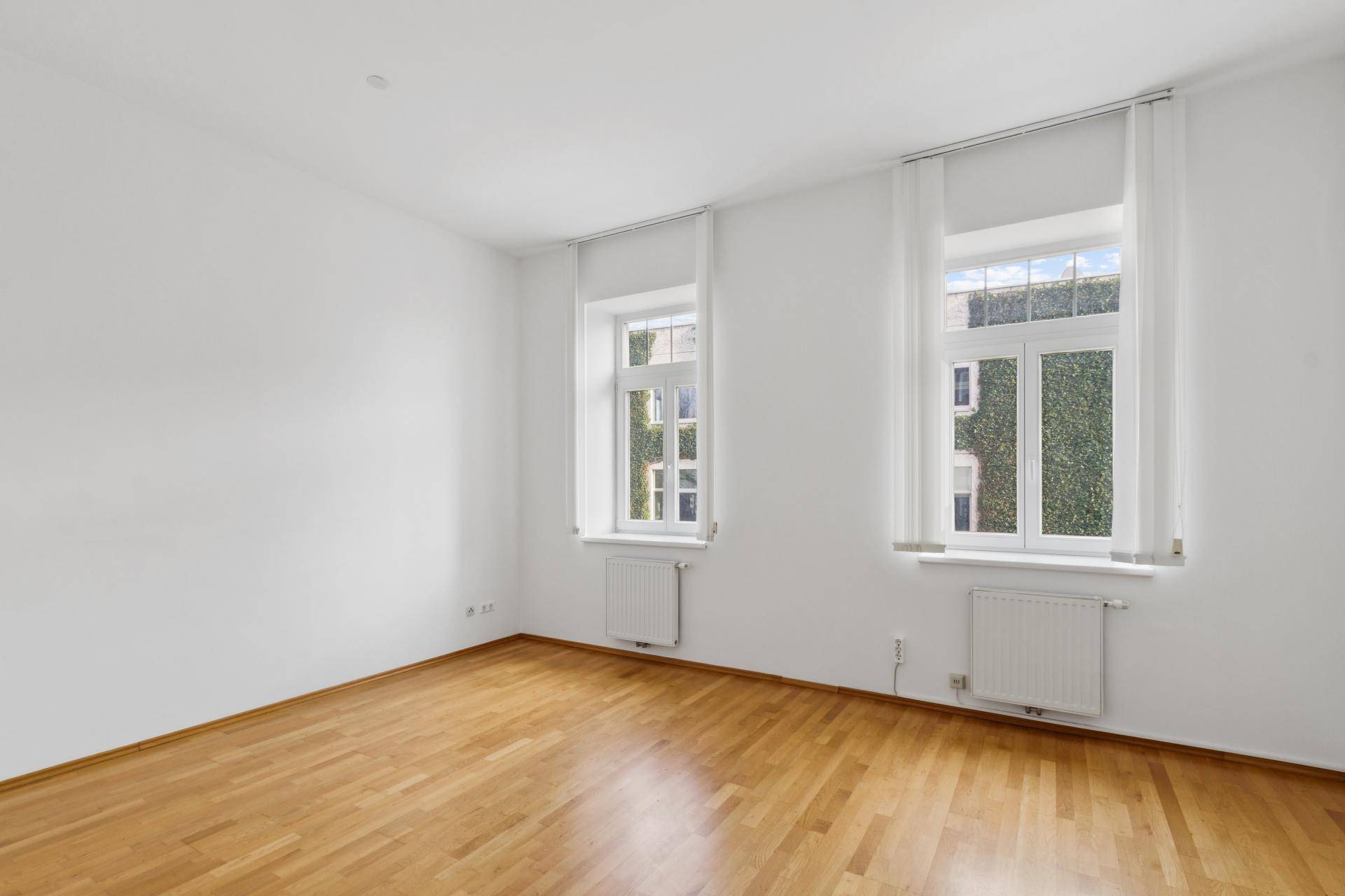 Leeres Zimmer mit Parkettboden und zwei Fenstern mit Blick ins Grüne.