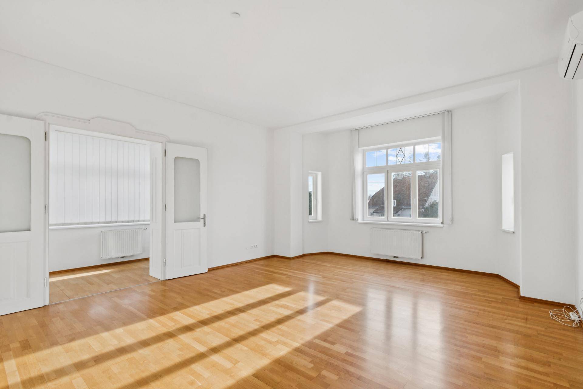 Helles Wohnzimmer mit Parkettboden, großen Fenstern und einer Klimaanlage.