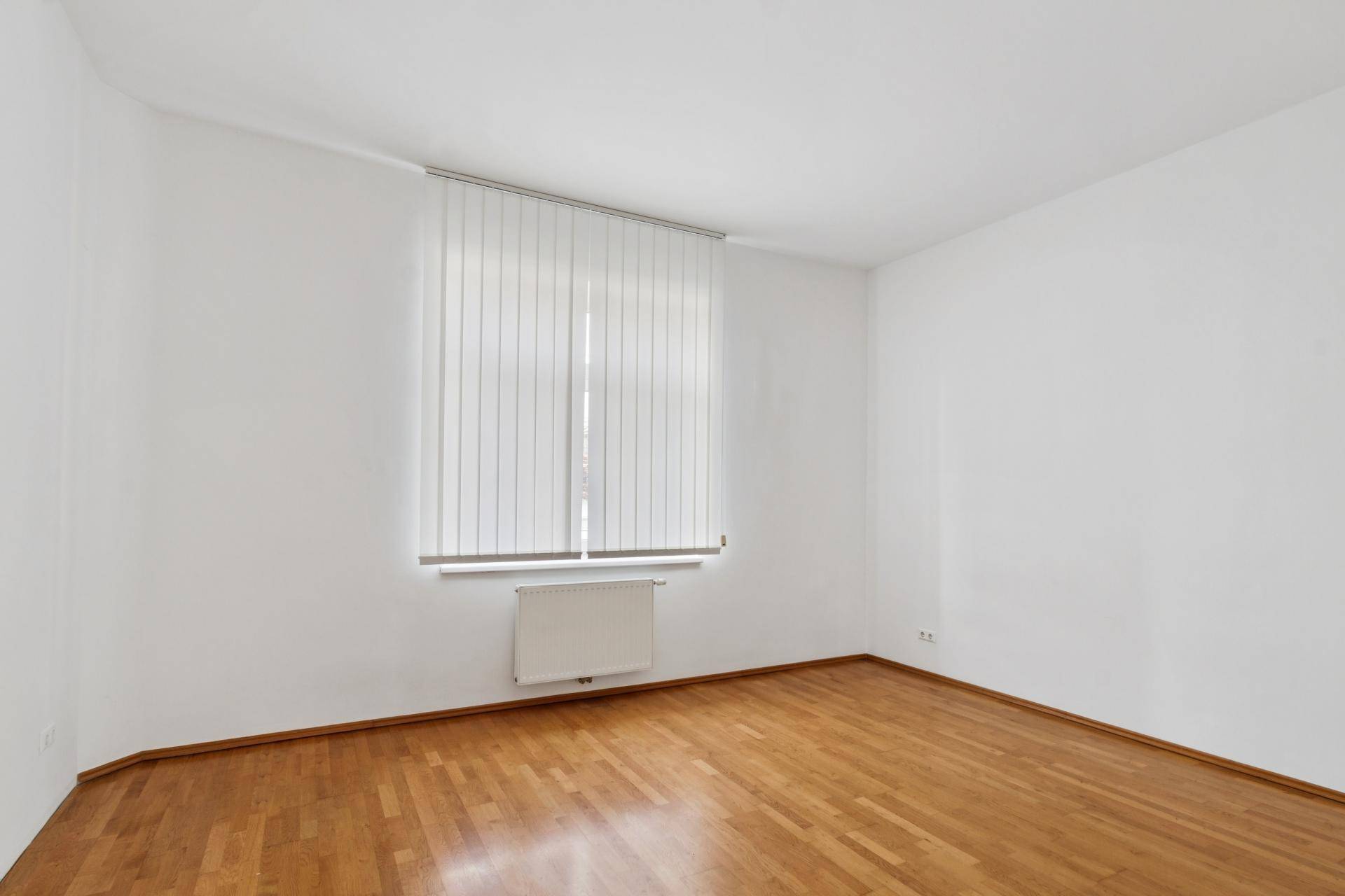 Leeres Zimmer mit Parkettboden und einem großen Fenster mit vertikalen Jalousien.