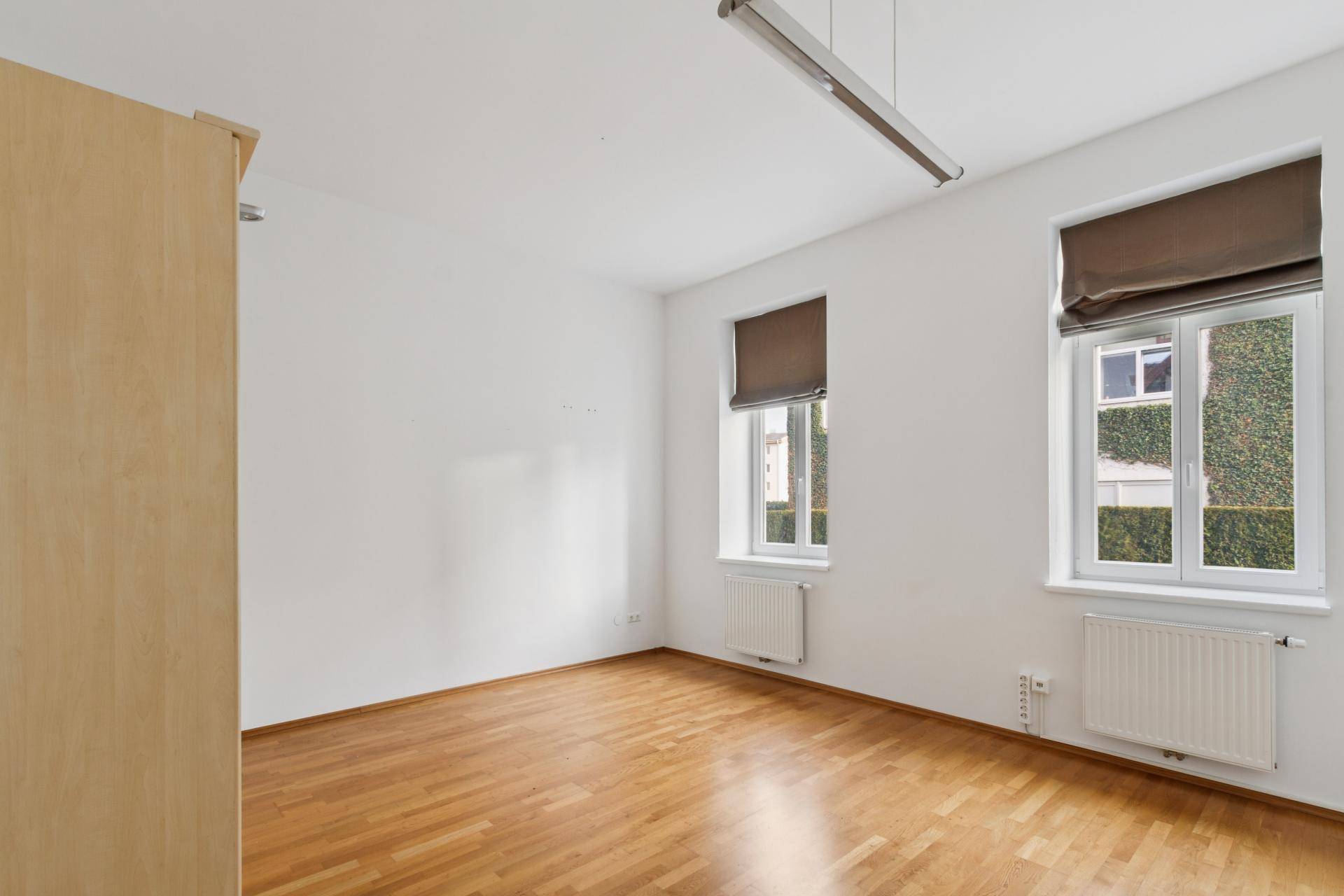Helles Zimmer mit Parkettboden und zwei Fenstern, die einen Blick ins Grüne bieten.