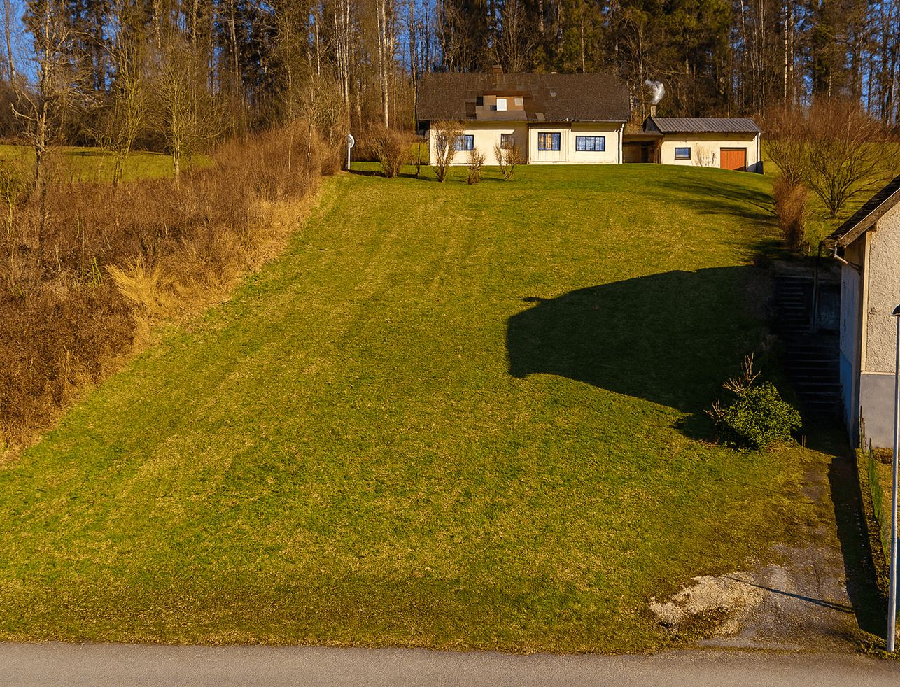 Haus auf einem grünen Hügel mit Wald im Hintergrund und einem weiteren Gebäude auf der rechten Seite.