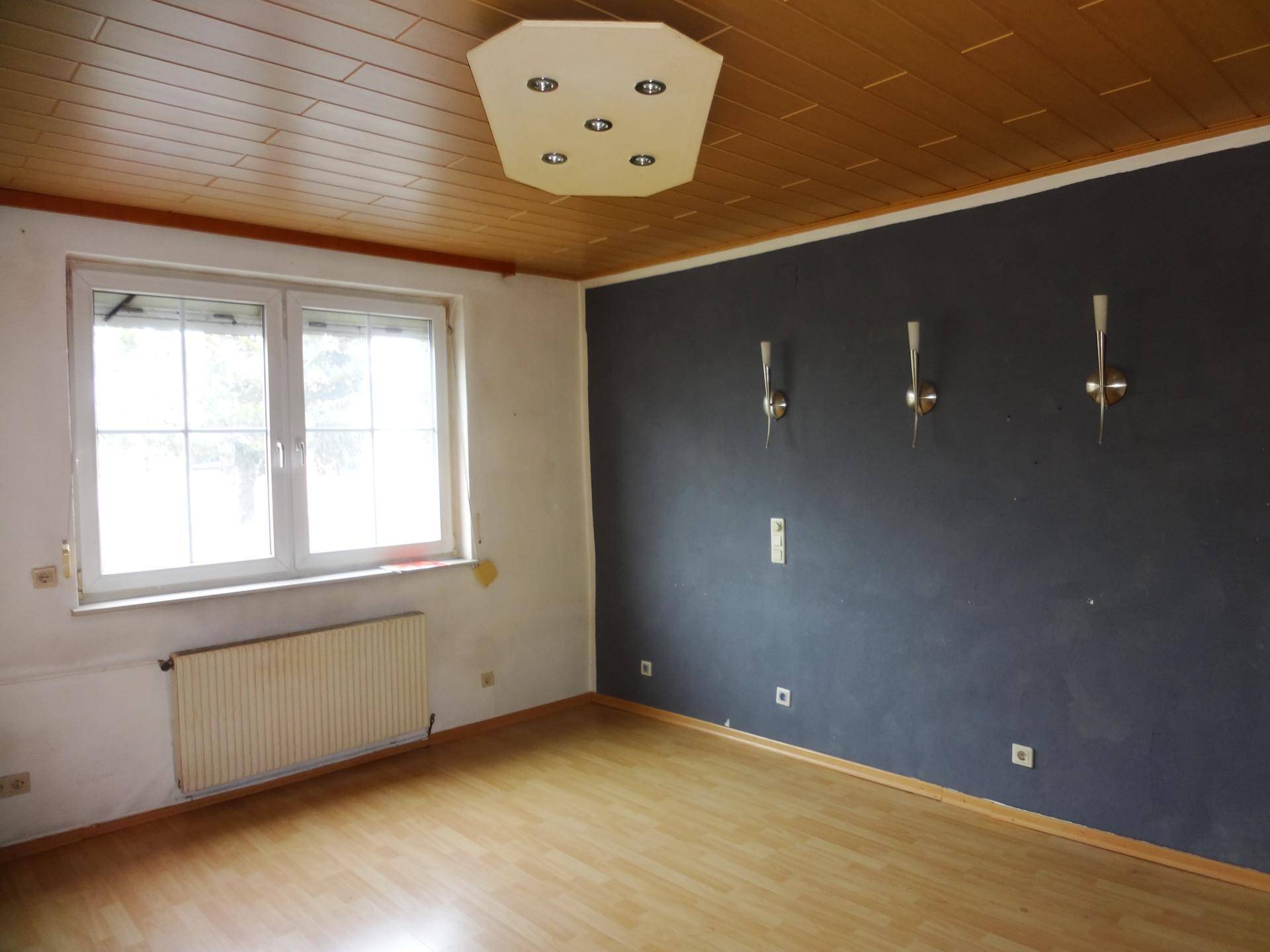 Zimmer mit dunkler Akzentwand, Holzpaneeldecke und Laminatboden, beleuchtet von einer Deckenleuchte und Wandlampen.