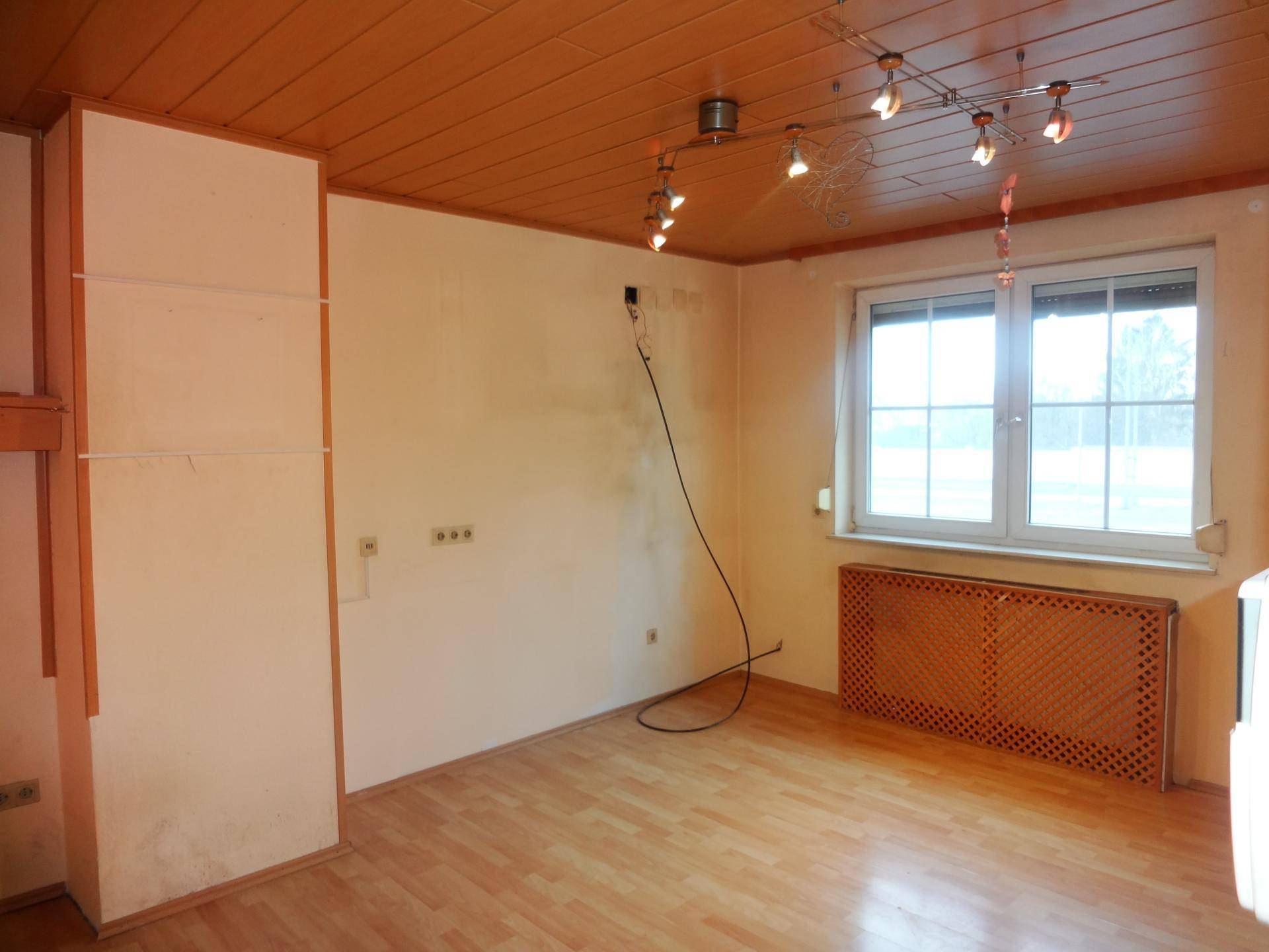 Helles Zimmer mit Holzpaneeldecke, Laminatboden und einem Heizkörper unter dem Fenster, in originalem Zustand.