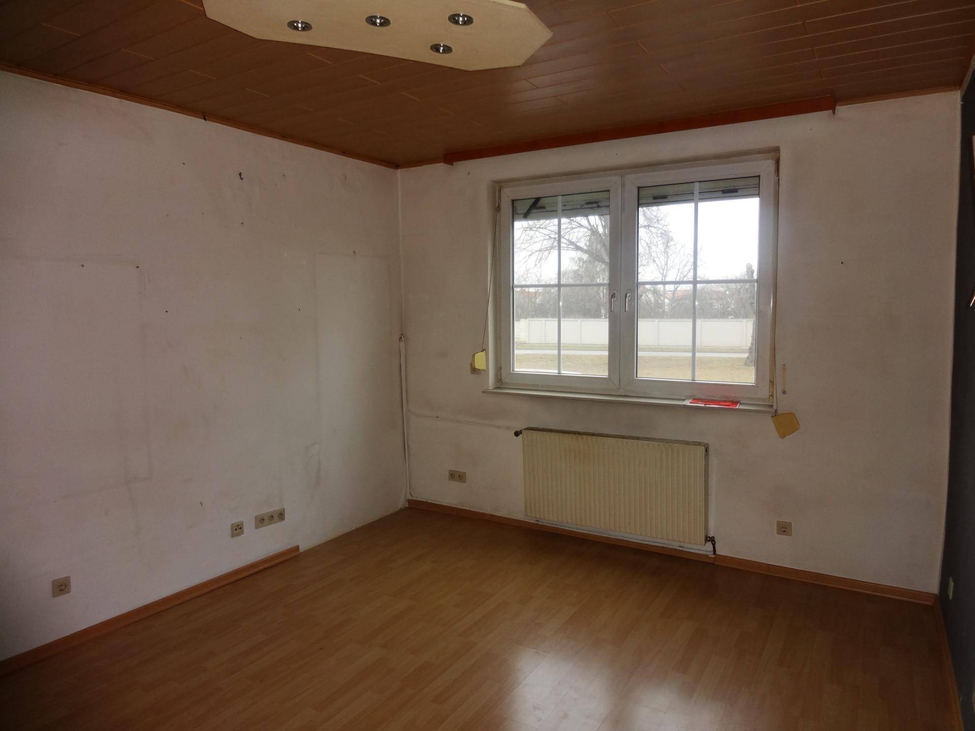 Leeres Zimmer mit Holzpaneeldecke, Laminatboden und einem Heizkörper unter dem Fenster, mit viel Tageslicht.
