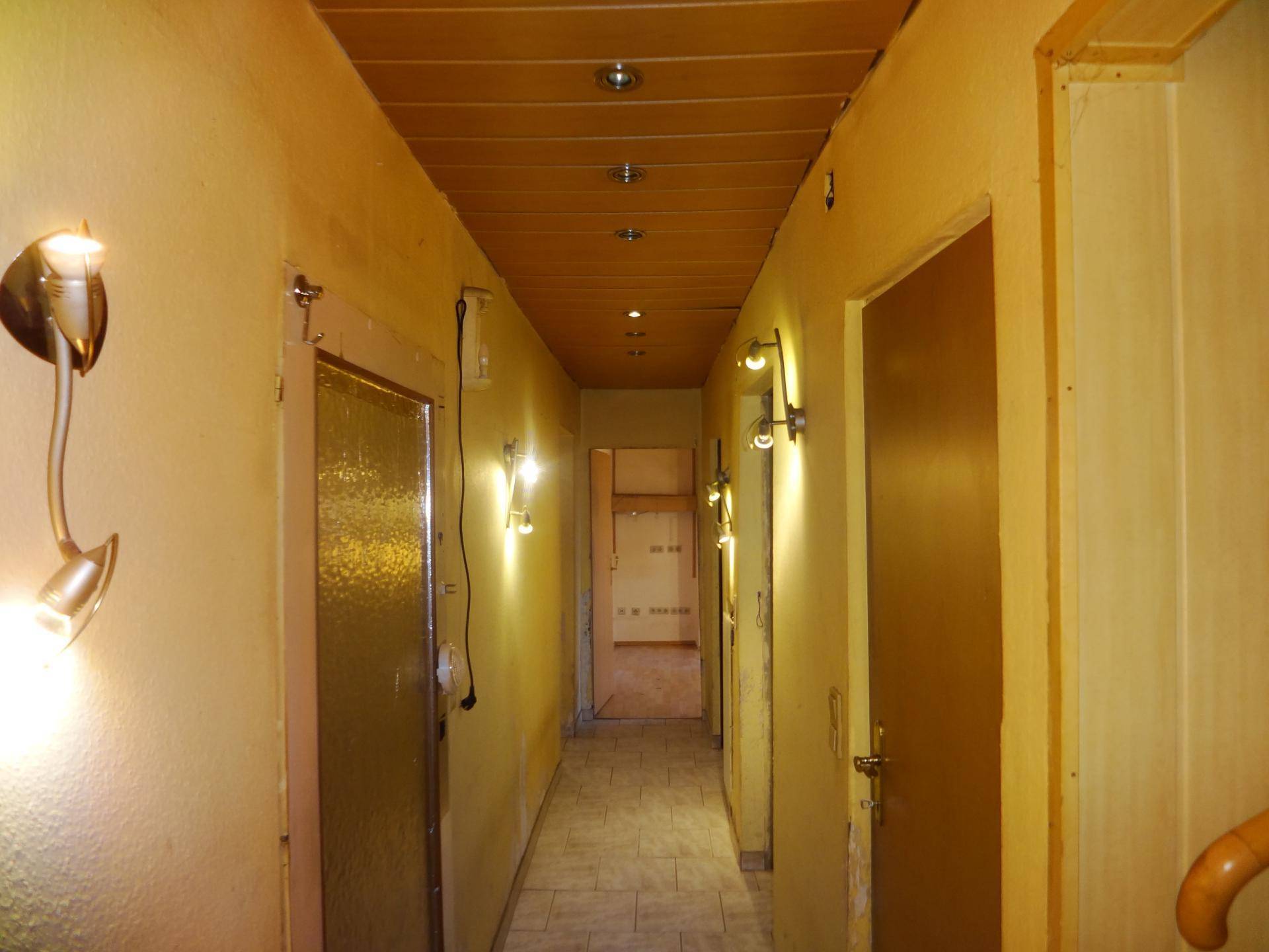 Langer Flur mit Holzpaneeldecke, Fliesenboden und mehreren Türen, beleuchtet von Wandleuchten.