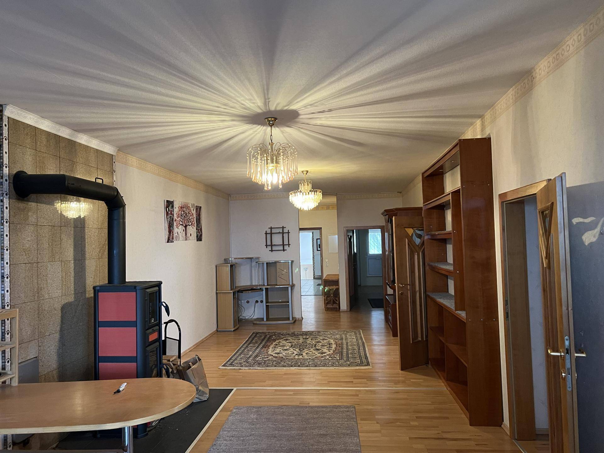 Geräumiges Wohnzimmer mit Holzboden, einem Kaminofen und traditionellen Möbeln unter Kronleuchtern.