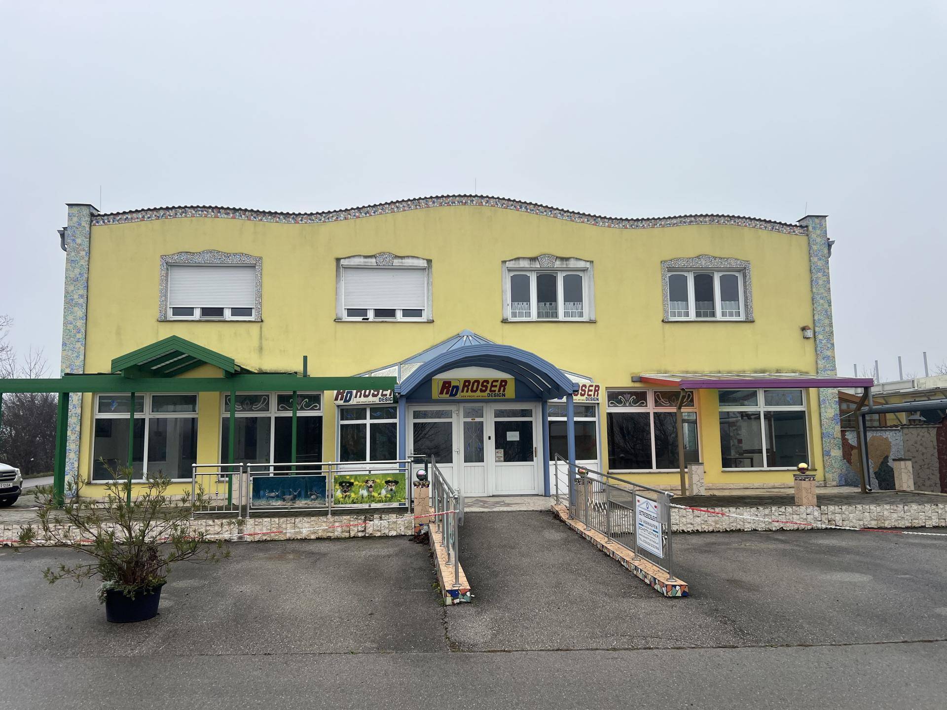 Gelbes Geschäftsgebäude mit mehreren Fenstern, Rollläden und einem Eingangsbereich mit Rampe für Barrierefreiheit.