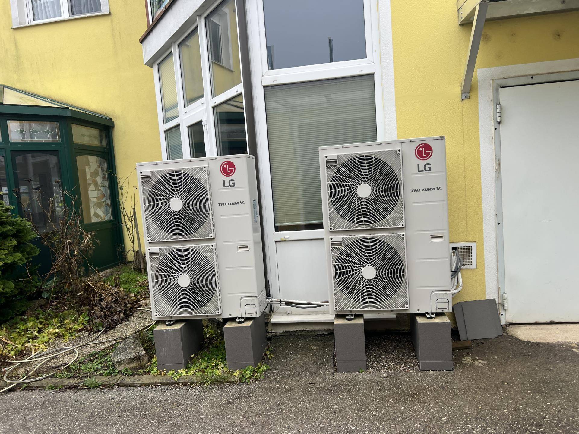 Zwei große Klimaanlagen-Außengeräte stehen vor einer gelben Gebäudefassade mit Fenstern.