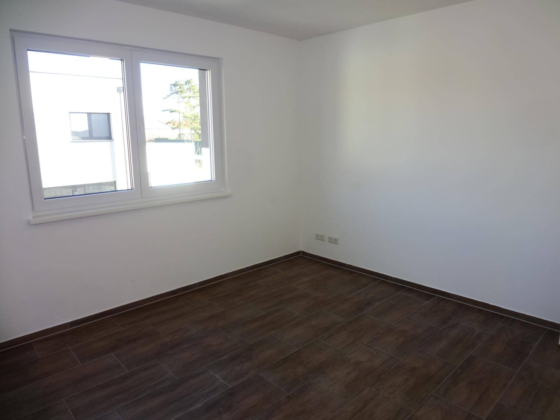 Leeres Schlafzimmer mit hellem Fenster und dunklem Fliesenboden in neuwertigem Zustand.
