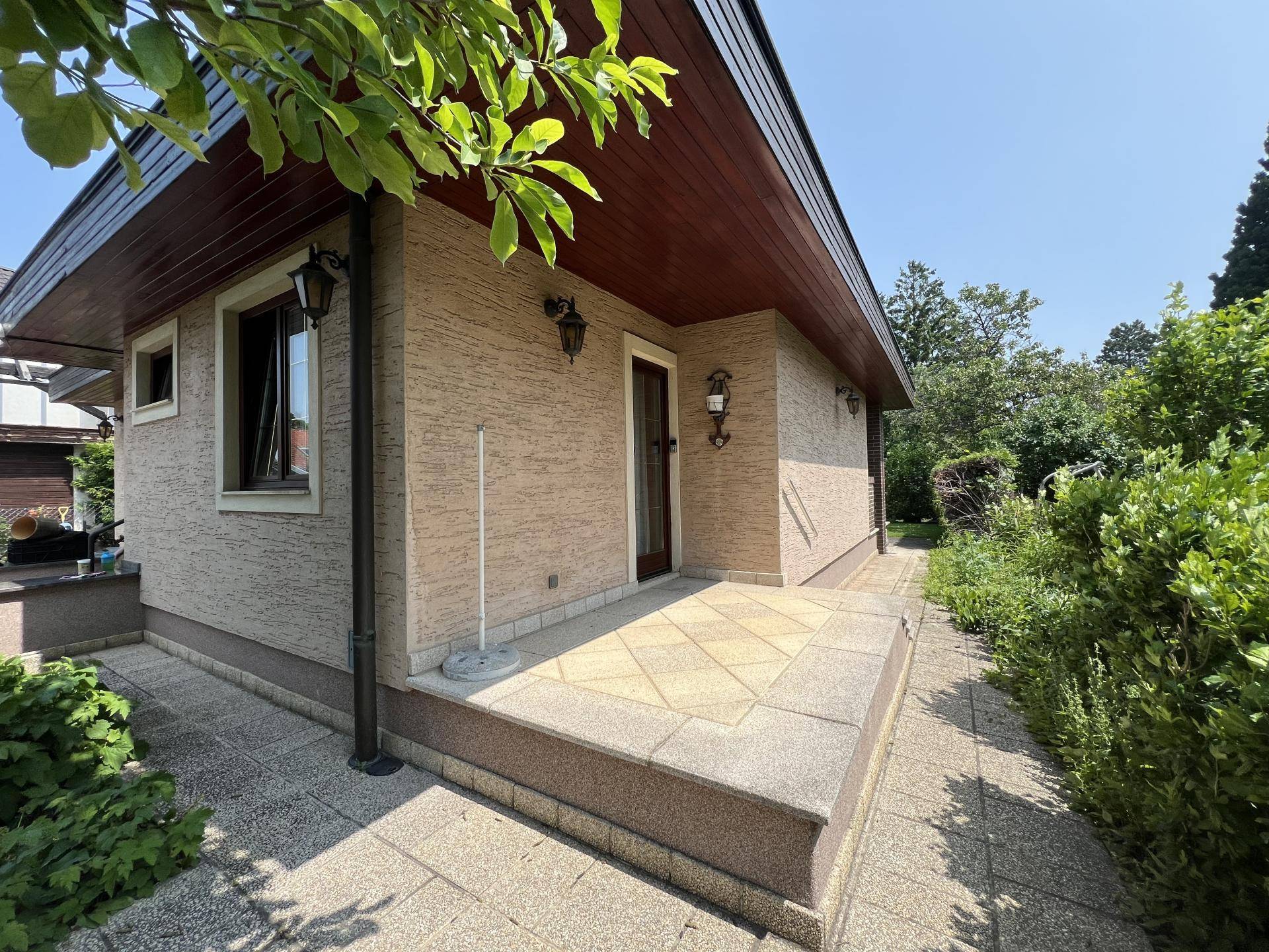 Seitliche Terrasse mit Fliesenboden und Zugang zum Garten, umgeben von grünen Büschen.
