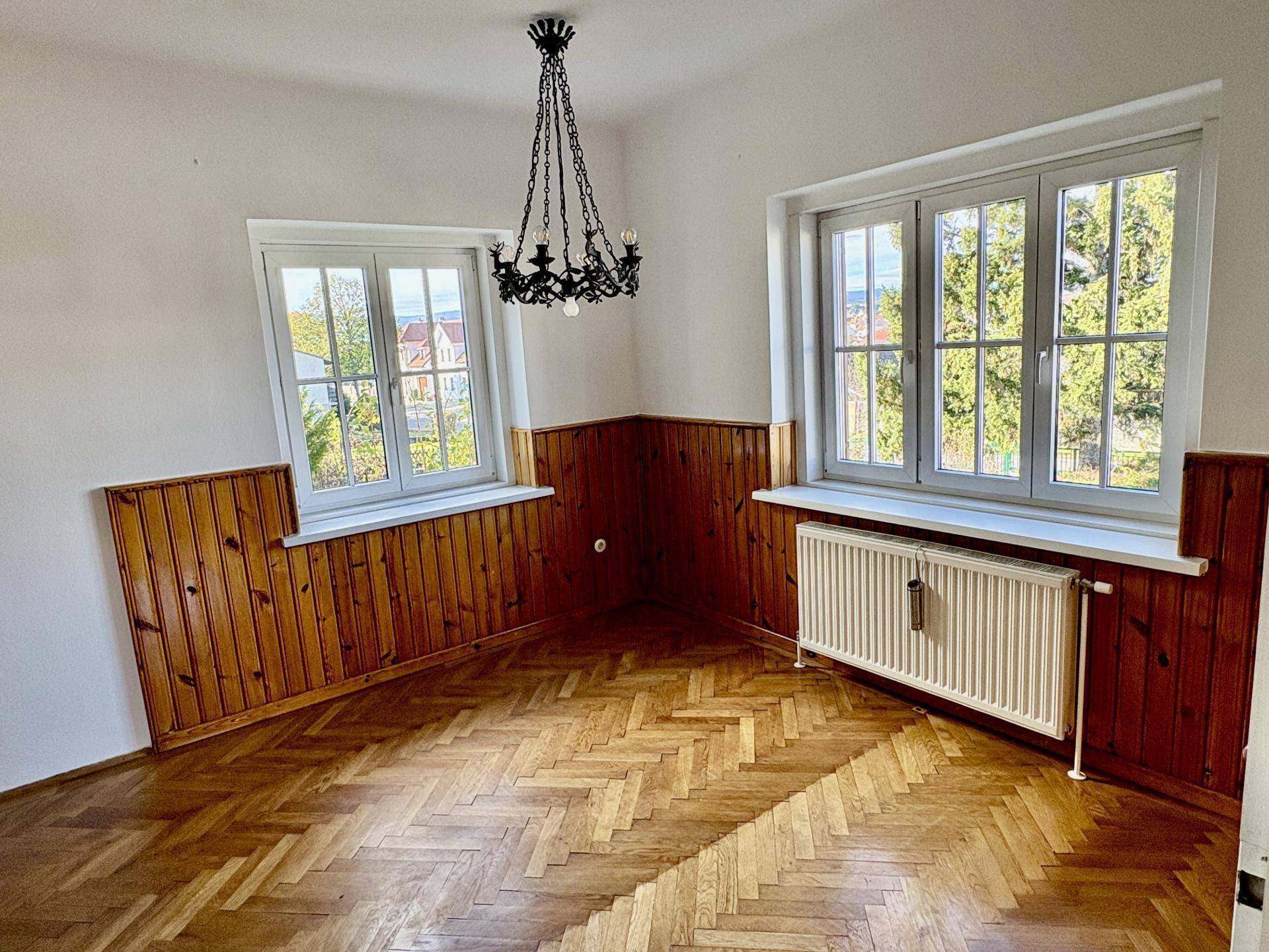 Leeres Zimmer mit Fischgrätparkett, Holzvertäfelung und zwei großen Fenstern, die viel Tageslicht hereinlassen.
