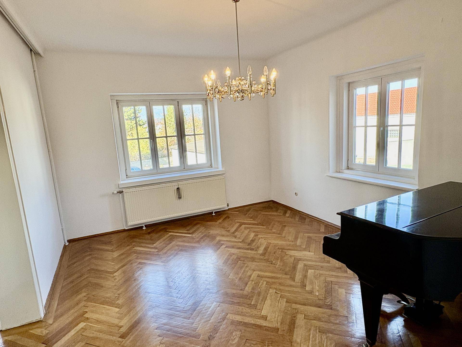 Geräumiges Zimmer mit glänzendem Fischgrätparkett, einem Flügel und einem Kronleuchter, hell durch große Fenster.