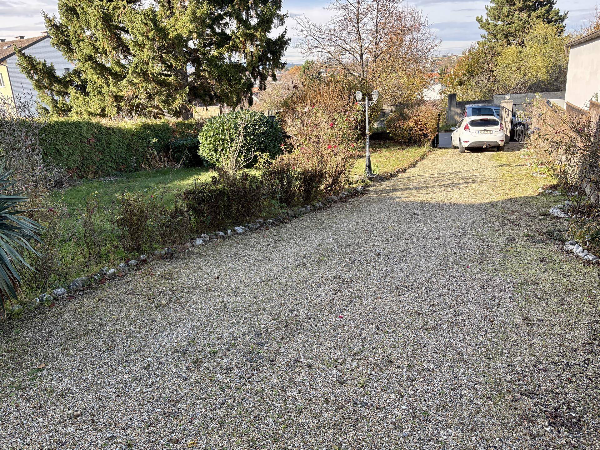 Großer Gartenbereich mit Kiesauffahrt und parkenden Autos, umgeben von Bäumen und Sträuchern.