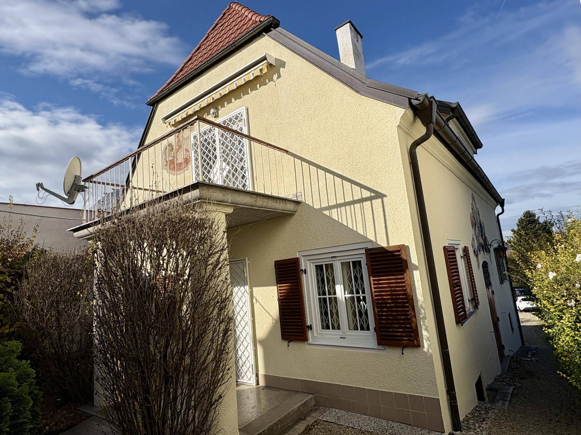 Gelbes Haus mit Balkon, braunen Fensterläden und einem Schornstein unter blauem Himmel, umgeben von Sträuchern.