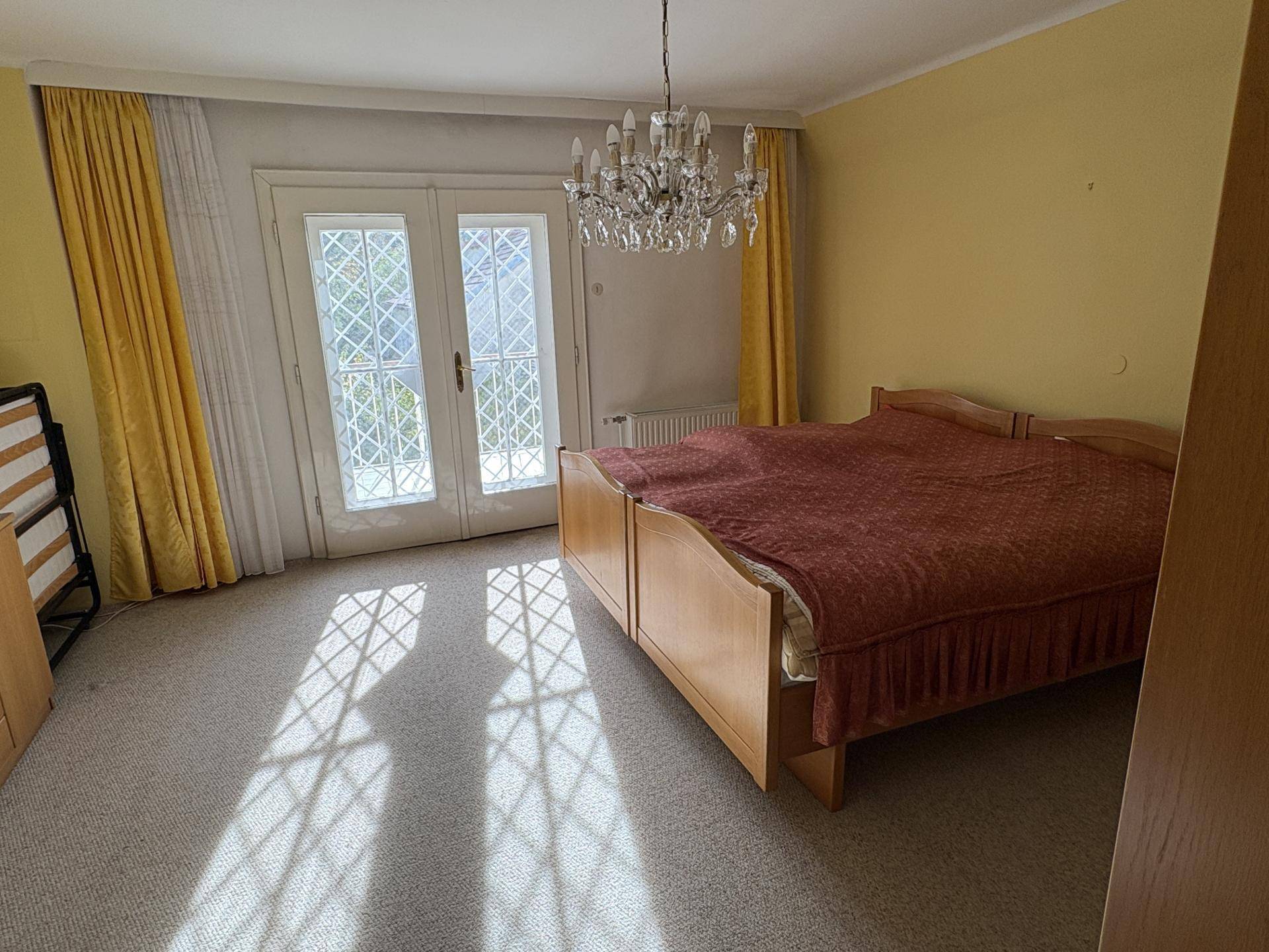 Schlafzimmer mit Teppichboden, einem Doppelbett und Balkonzugang durch große Glastüren, hell beleuchtet.