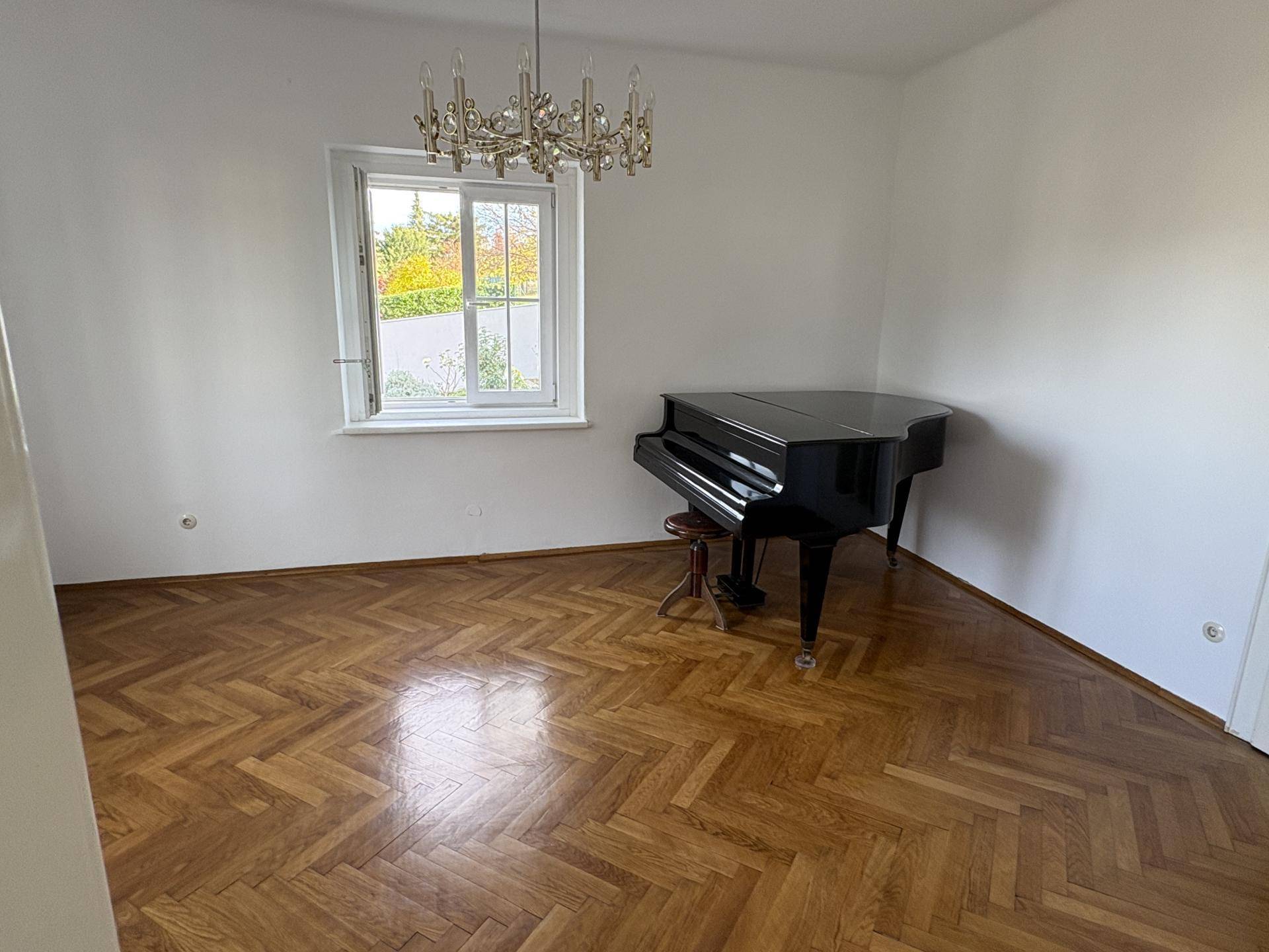 Helles Zimmer mit Fischgrätparkett und einem schwarzen Flügel, beleuchtet durch ein großes Fenster mit Ausblick.