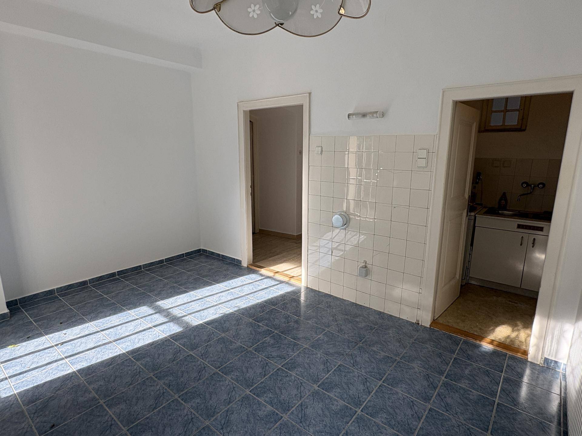Einfacher Raum mit blau gefliestem Boden und einer kleinen Küchenzeile in einem angrenzenden Bereich, hell beleuchtet.