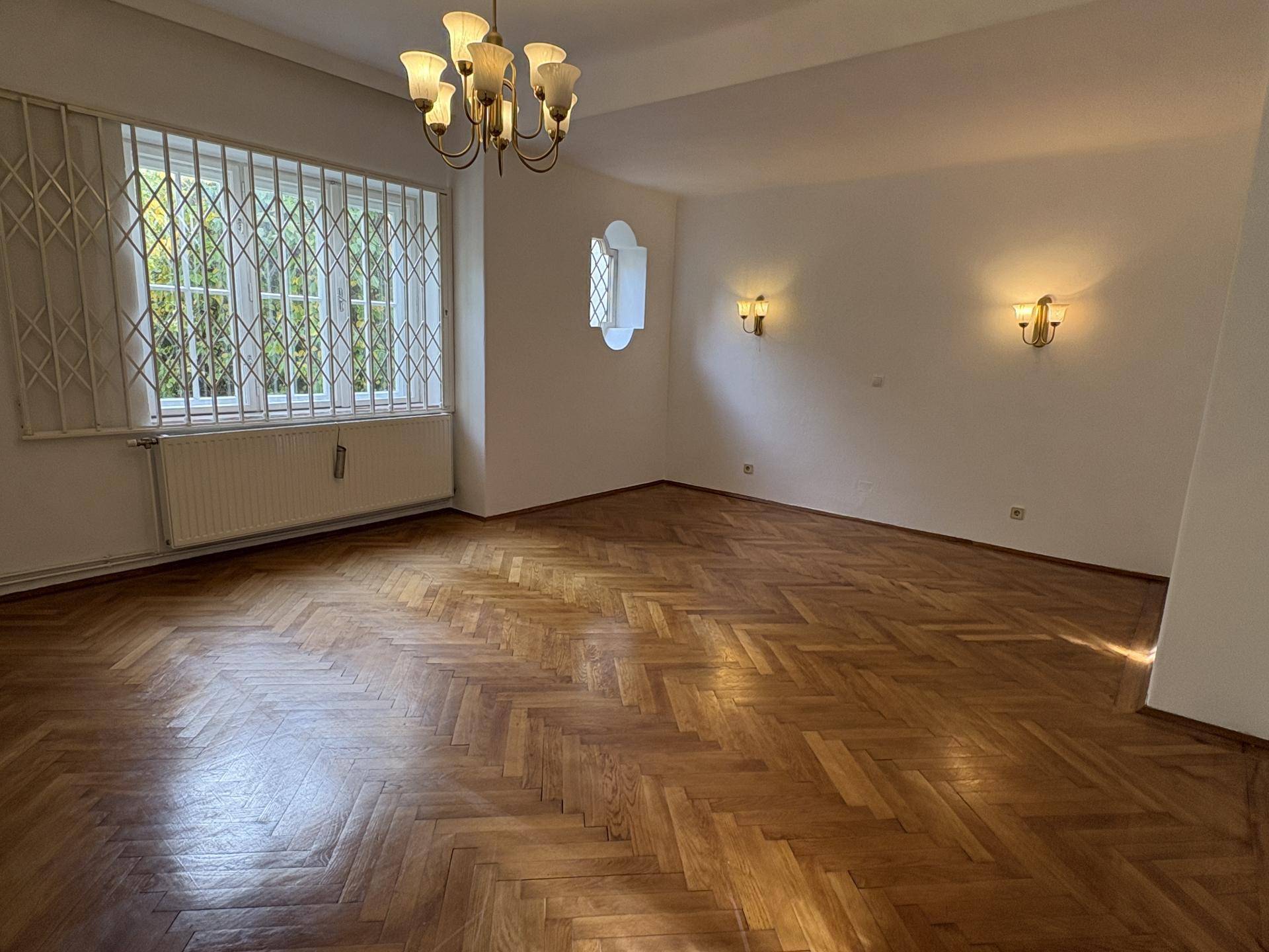 Geräumiger Wohnbereich mit Fischgrätparkett, einem Fenster mit Gitter und einem eleganten Kronleuchter an der Decke.