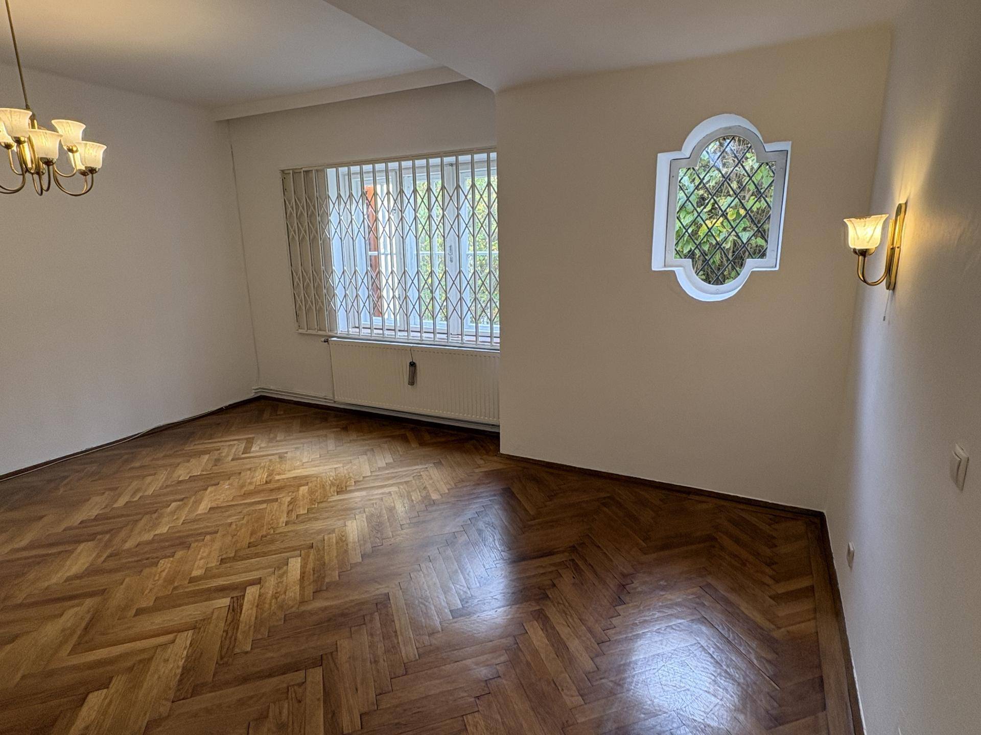 Wohnraum mit Fischgrätparkett, einem Fenster mit Sicherheitsgittern und dekorativen Wandleuchten, die eine gemütliche Atmosphäre schaffen.