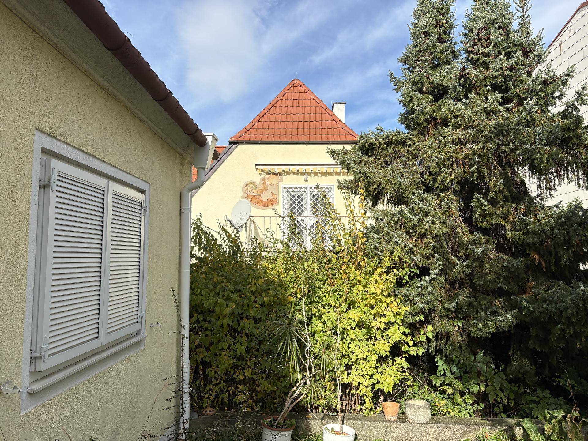 Seitenansicht eines gelben Hauses mit grüner Bepflanzung und einem Fenster mit Rollladen unter blauem Himmel.