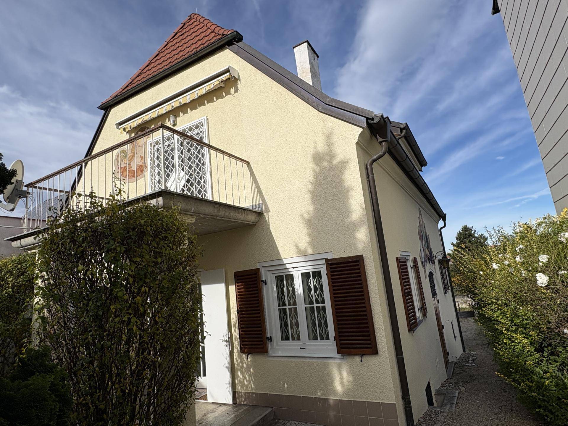 Gelbes Haus mit Balkon und braunen Fensterläden, umgeben von herbstlicher Vegetation unter blauem Himmel.