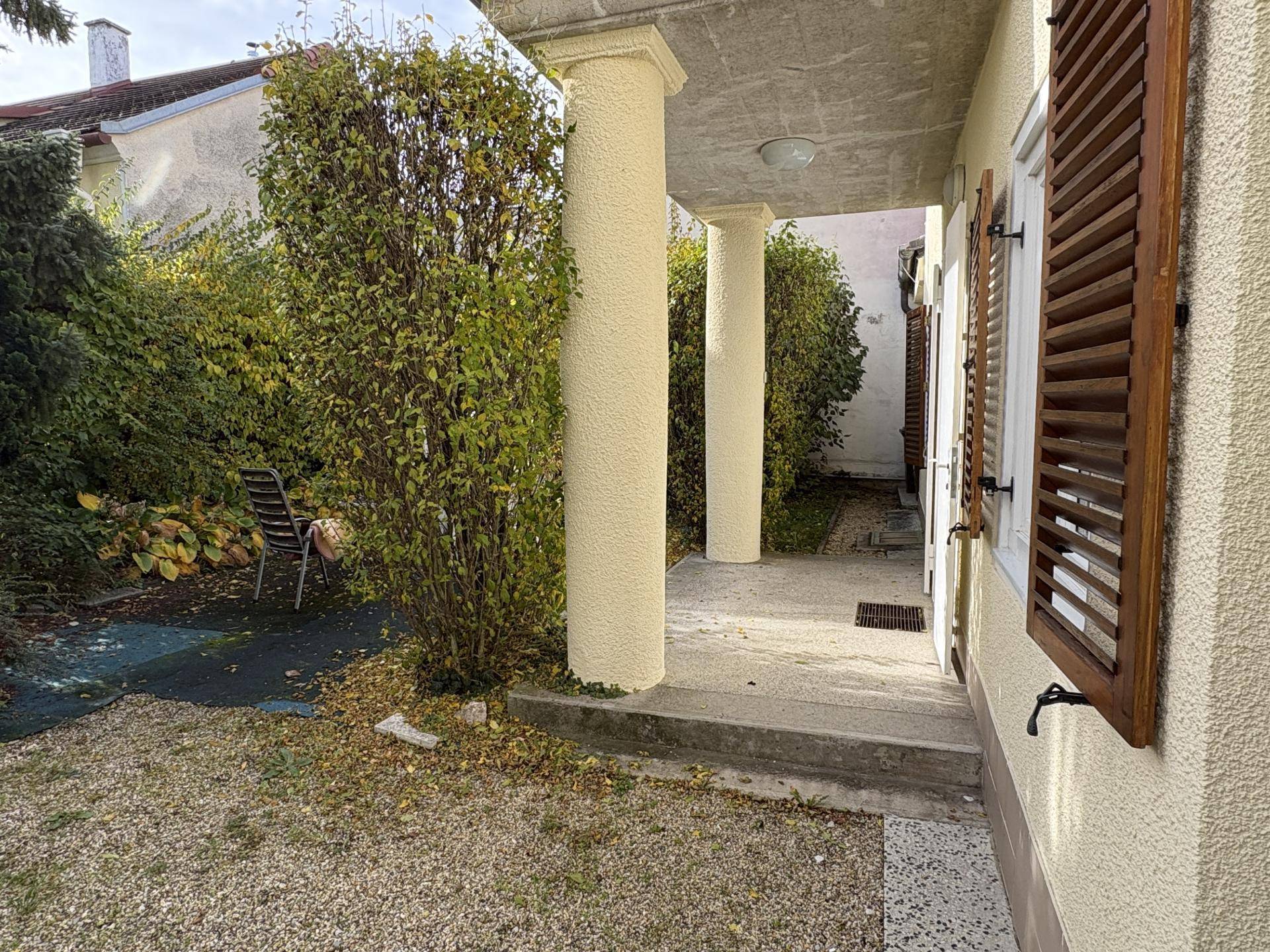 Überdachter Eingangsbereich mit Säulen und einem kleinen Gartenbereich, umgeben von Hecken und Sträuchern.