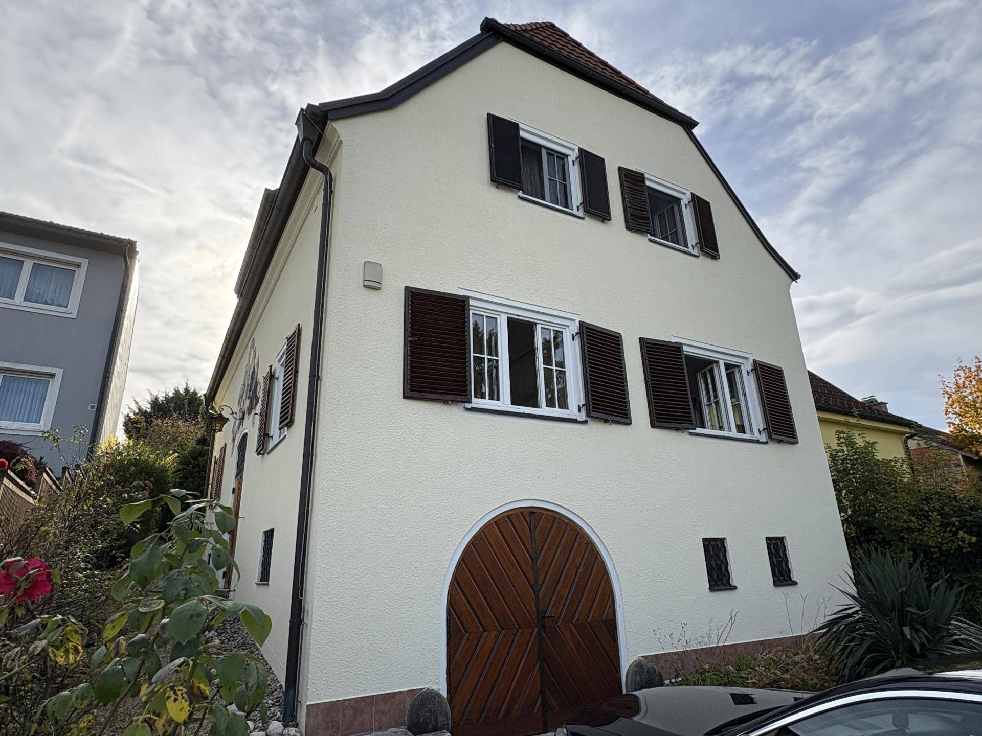 Weißes Haus mit braunen Fensterläden und einem großen Holztor, umgeben von herbstlicher Bepflanzung.