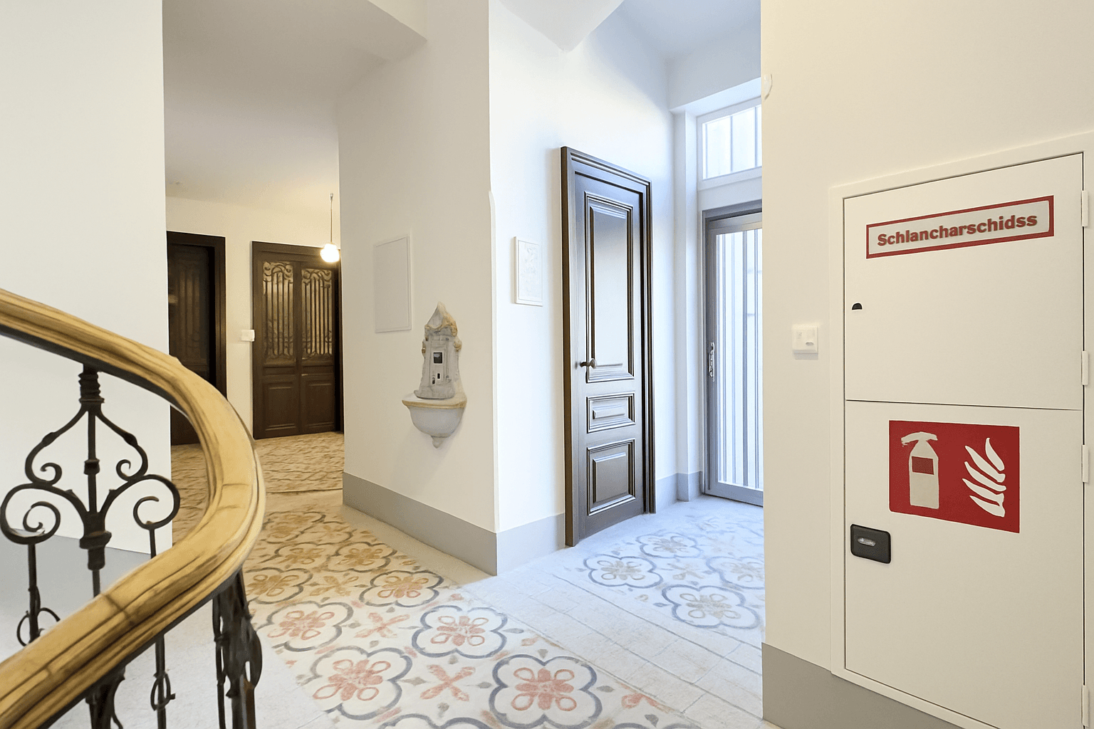 Historischer Treppenhausbereich mit kunstvollem Geländer, Fliesenboden und Brandschutzschrank.