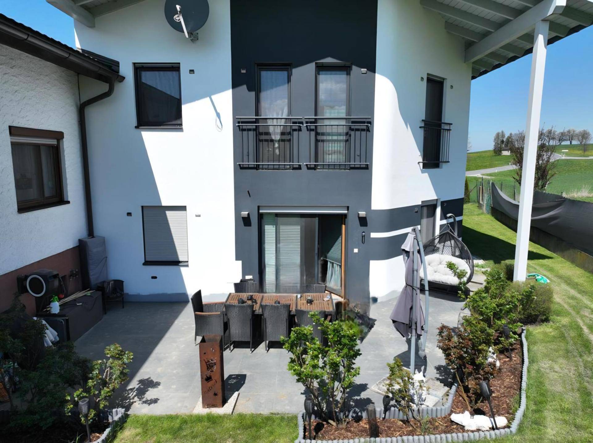 Moderne Hausfassade mit großer Terrasse, Gartenmöbeln und Blick ins Grüne.