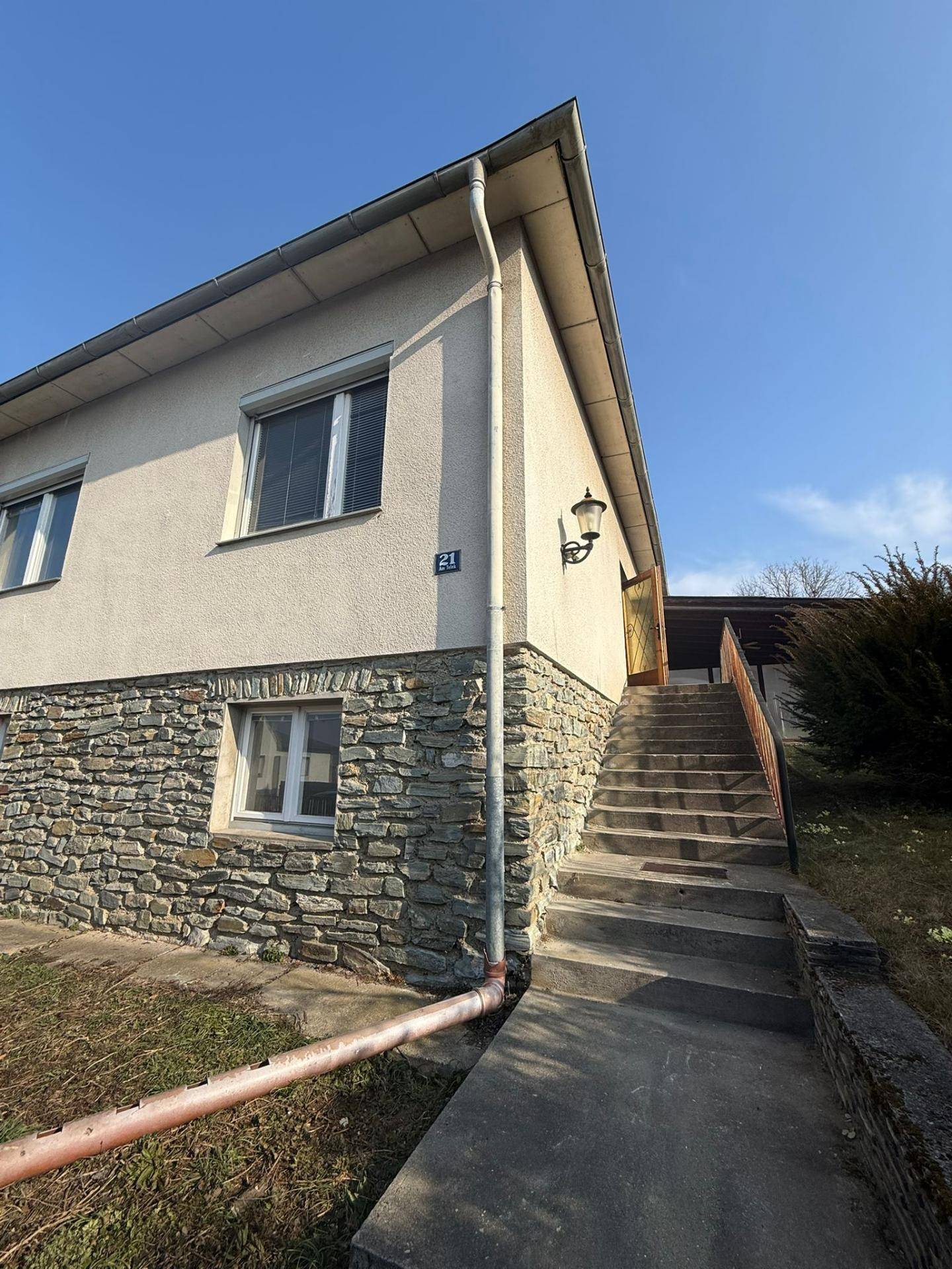 Seitenansicht des Hauses mit Außentreppe zum Eingang und Steinfassade im Sonnenlicht.