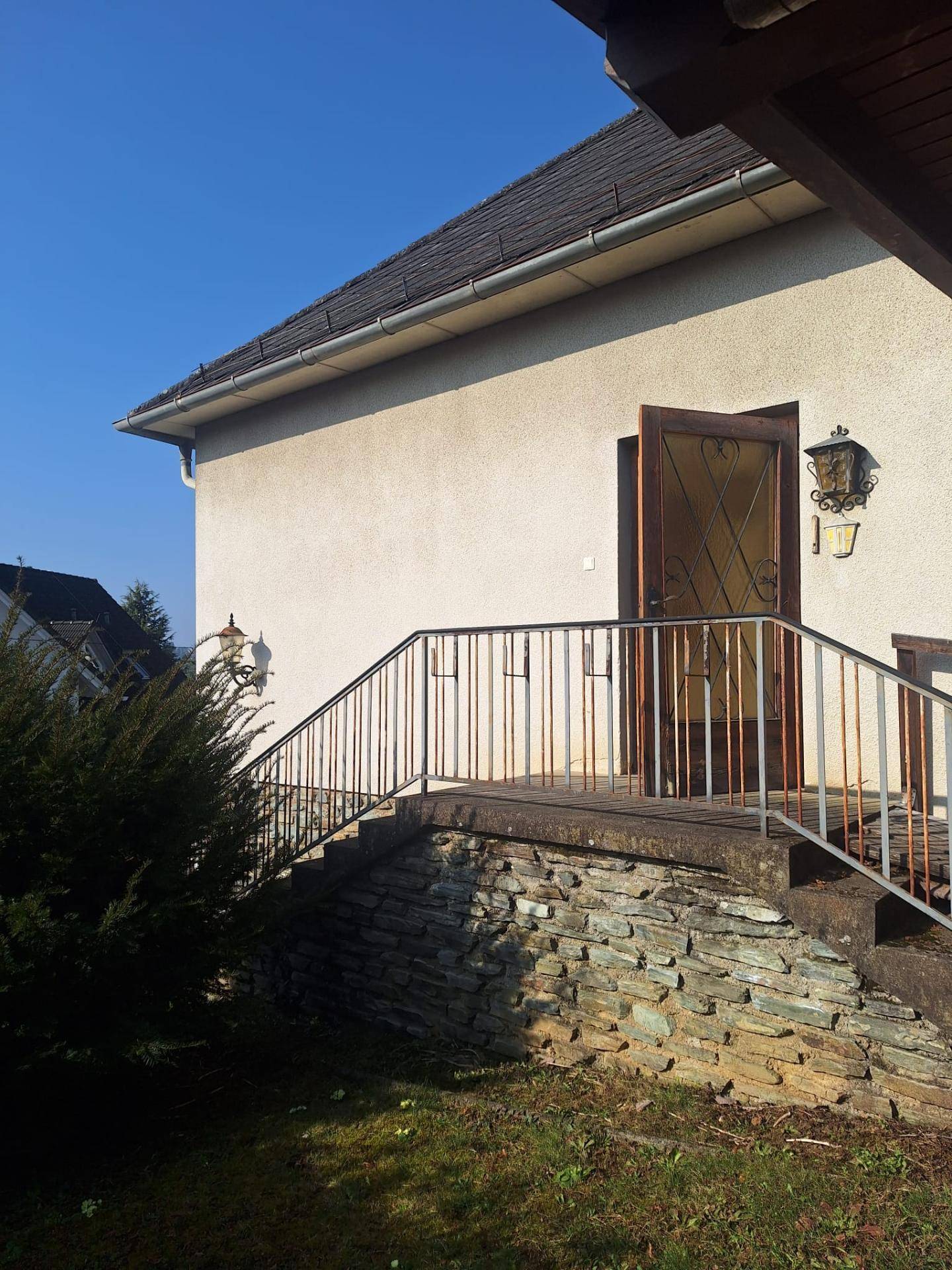 Eingangsbereich des Hauses mit Außentreppe, Geländer und Laterne, umgeben von Grün.