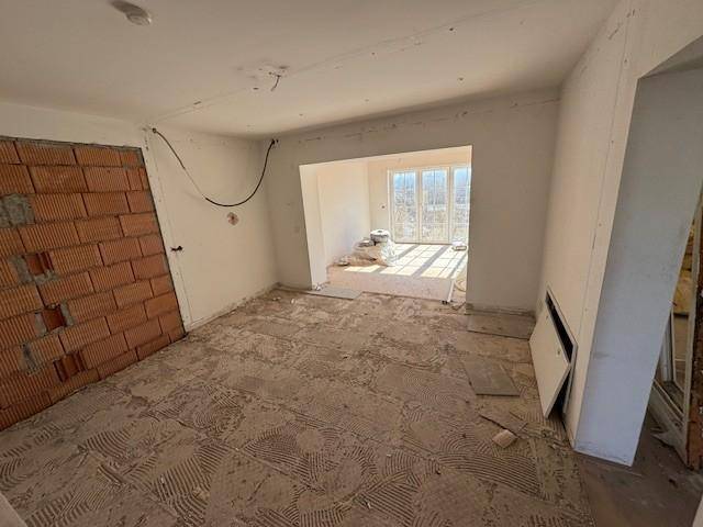 Innenraum im Rohbau mit unverputzter Ziegelwand und Blick in einen hellen angrenzenden Raum.