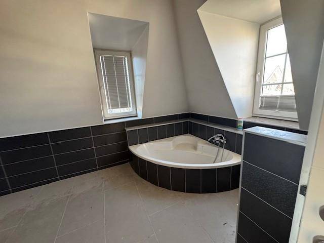 Modernes Badezimmer im Dachgeschoss mit Eckbadewanne, dunklen Fliesen und Fenster.