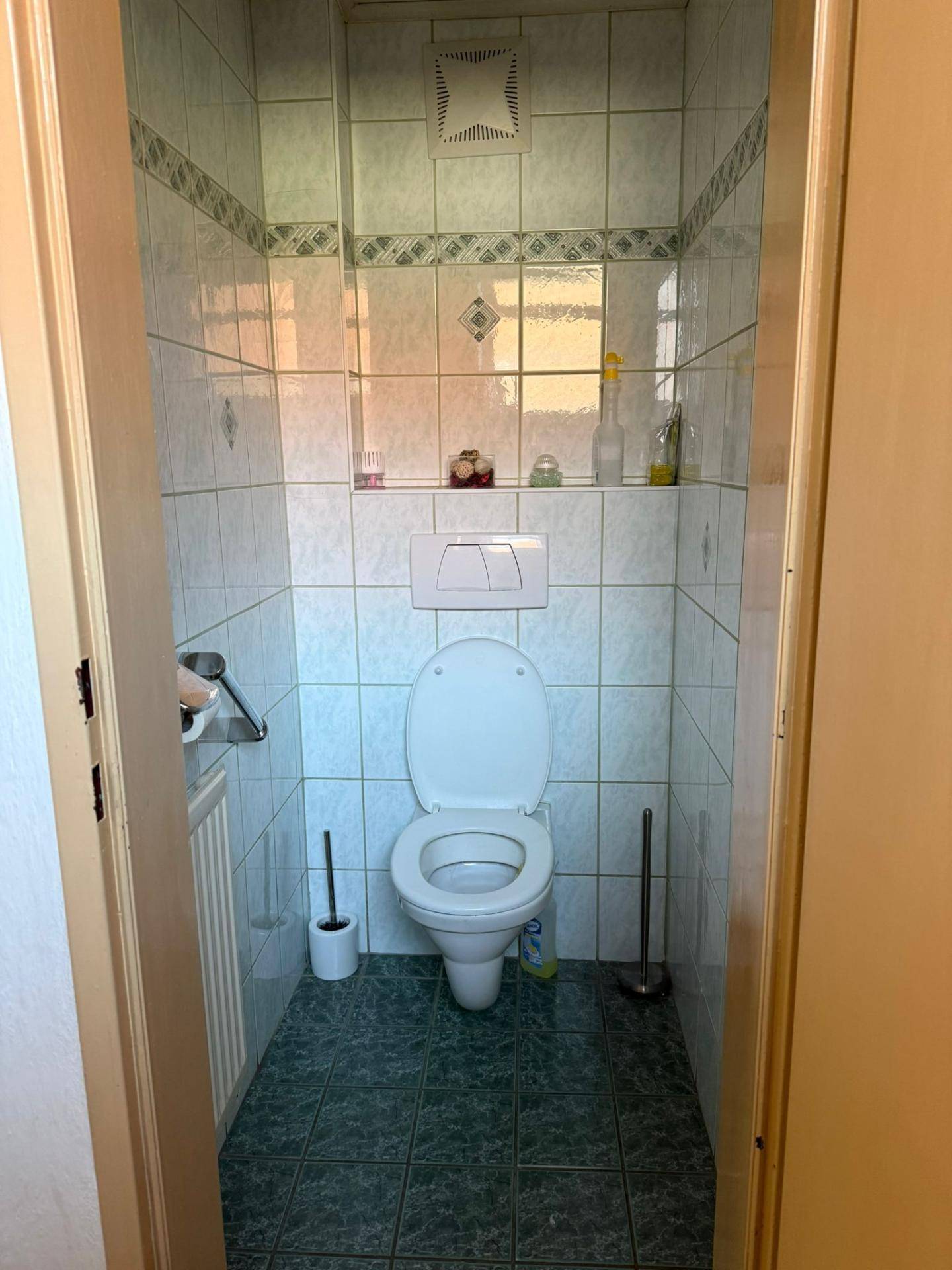 Separate Toilette mit Wand-WC, kleinem Waschbecken und grünen Bodenfliesen.