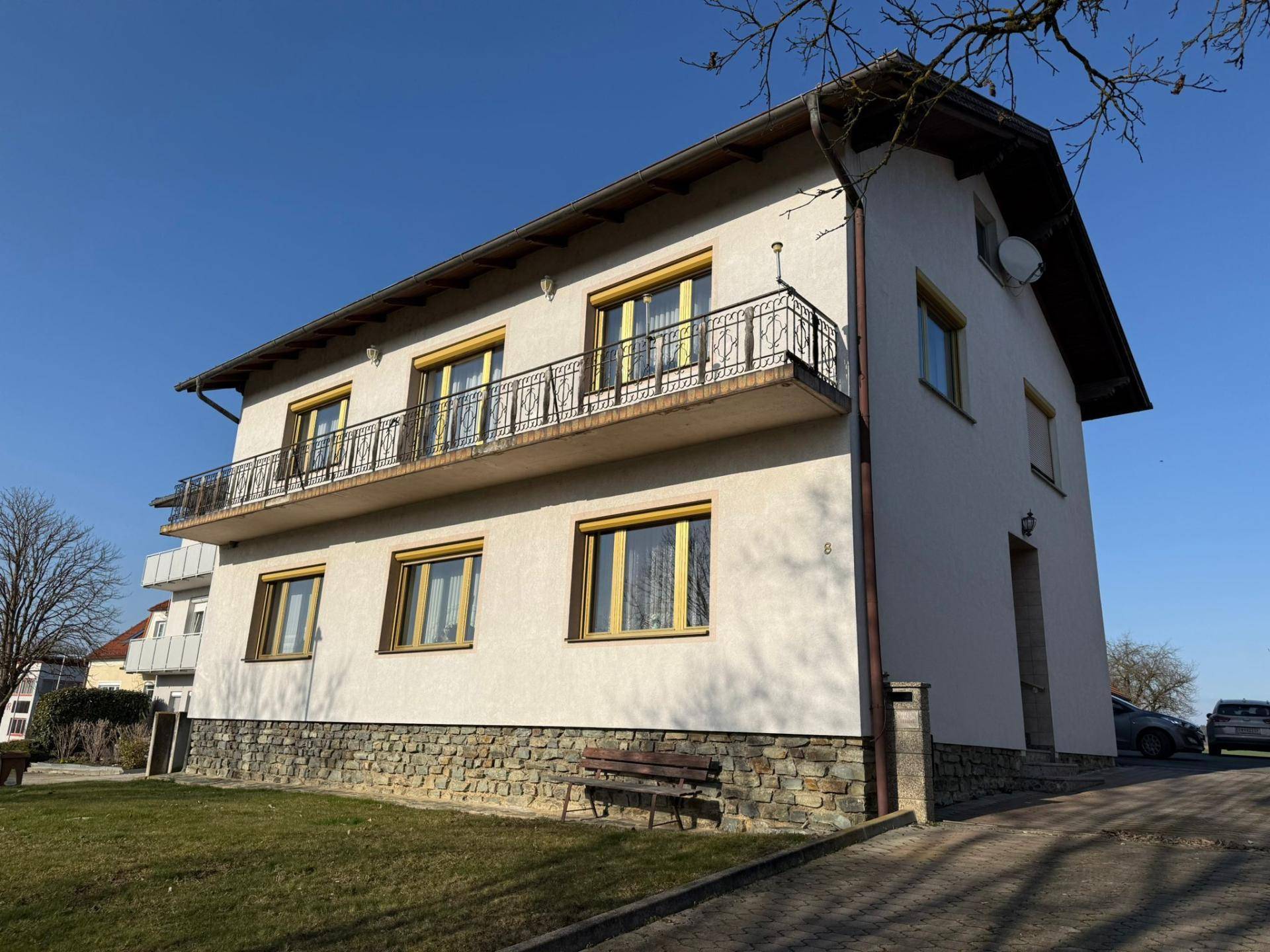 Einfamilienhaus mit Balkon, Steinsockel und gepflegtem Vorgarten unter blauem Himmel.