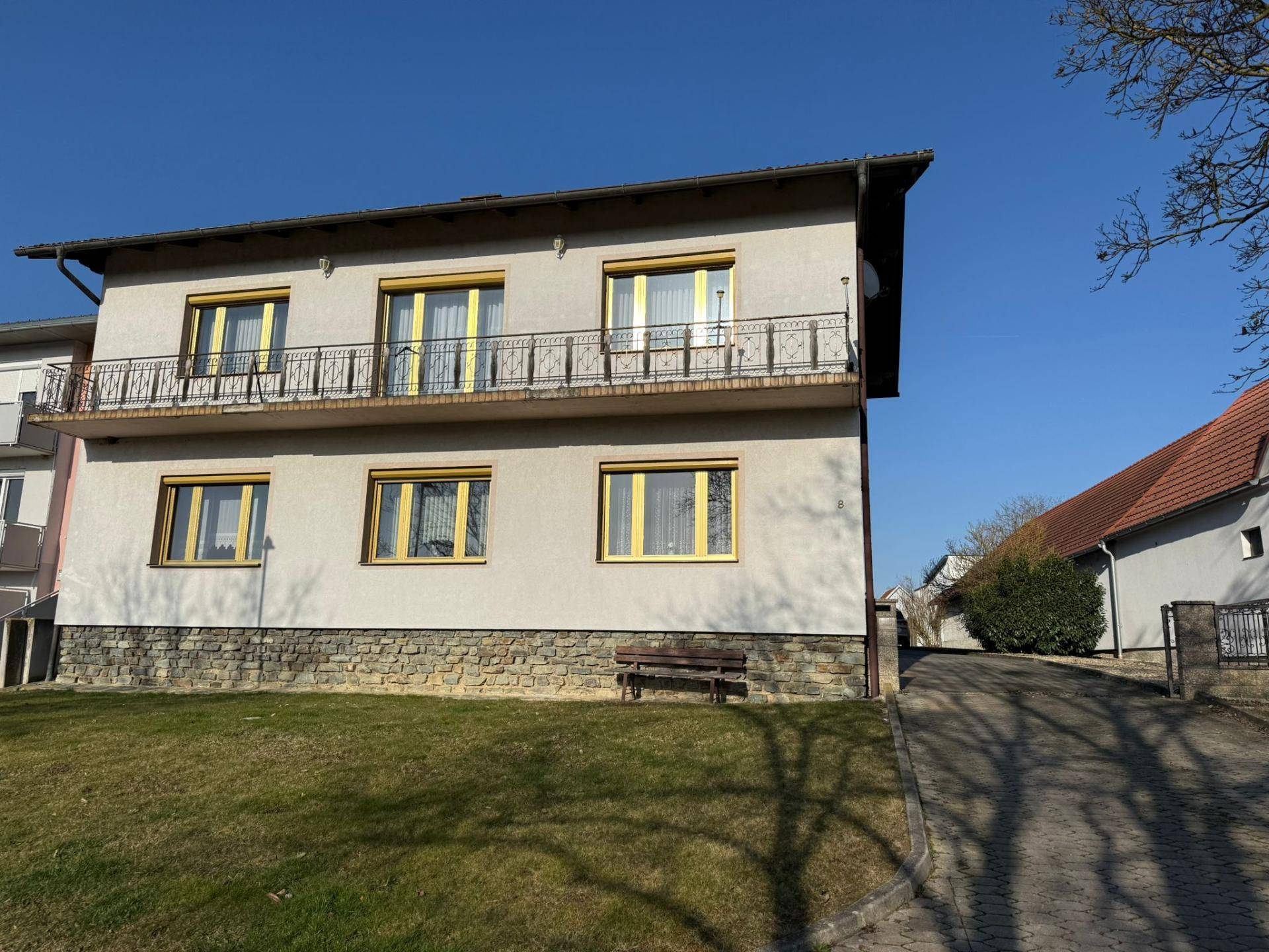 Einfamilienhaus mit Balkon, Steinsockel und gepflegtem Vorgarten unter blauem Himmel.