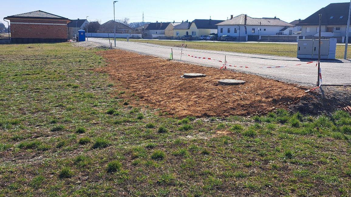 Grundstück mit vorbereitetem Bereich neben einer Straße, sichtbar sind Kanaldeckel und Markierungen.