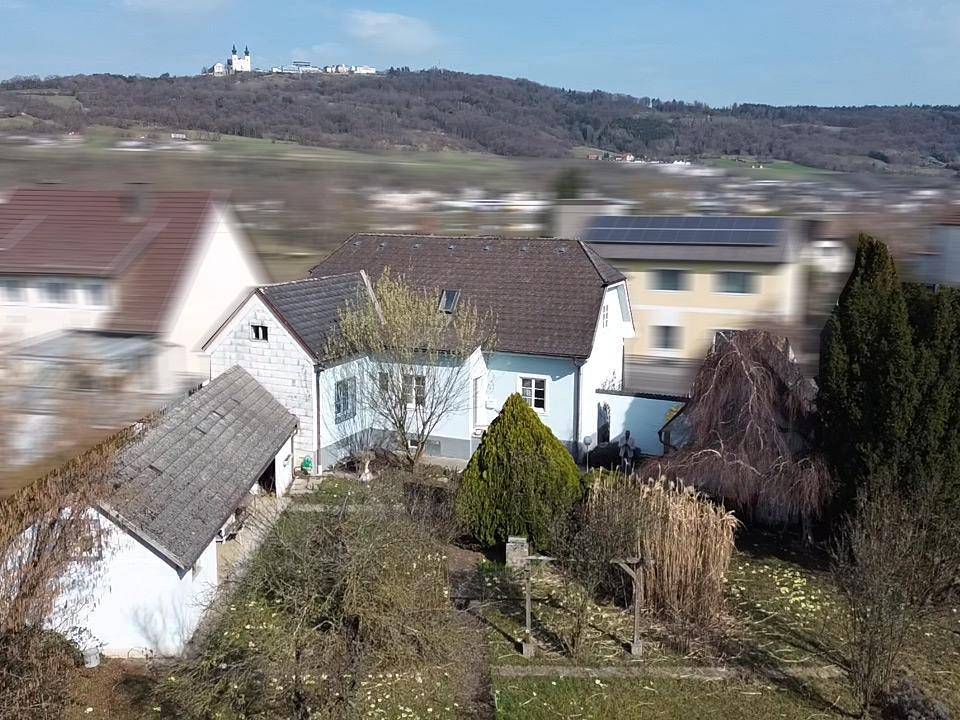 Luftaufnahme des Anwesens mit Garten, Nebengebäude und Fernblick auf eine Anhöhe.