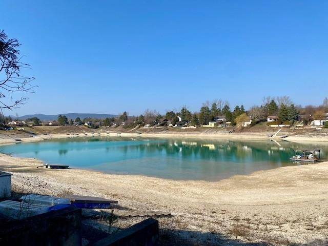 Weitläufiger Blick auf einen See mit flachem Ufer und Häusern im Hintergrund unter blauem Himmel.