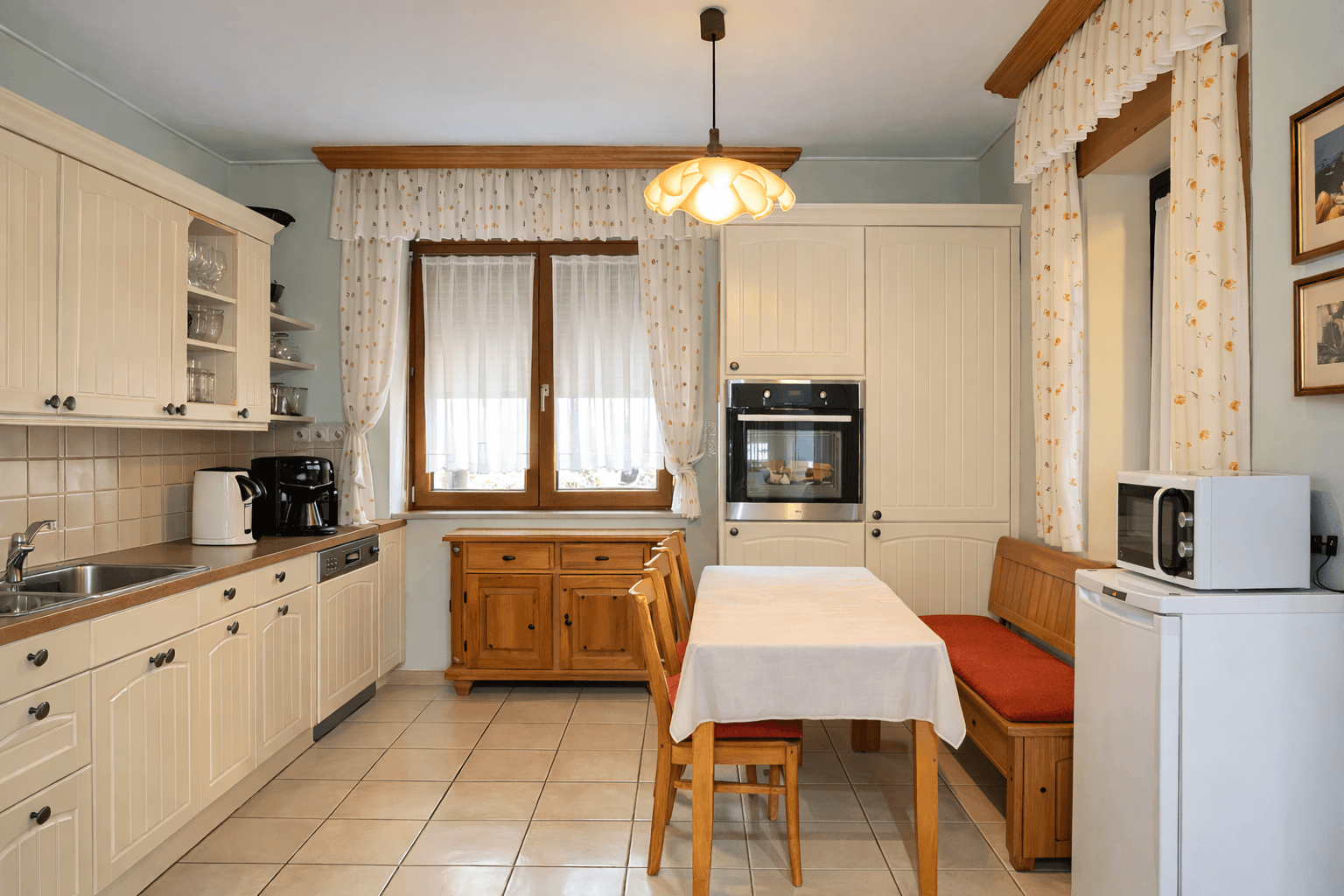 Gemütliche Wohnküche mit hellen Holzschränken, Esstisch und Fenster mit Vorhängen.