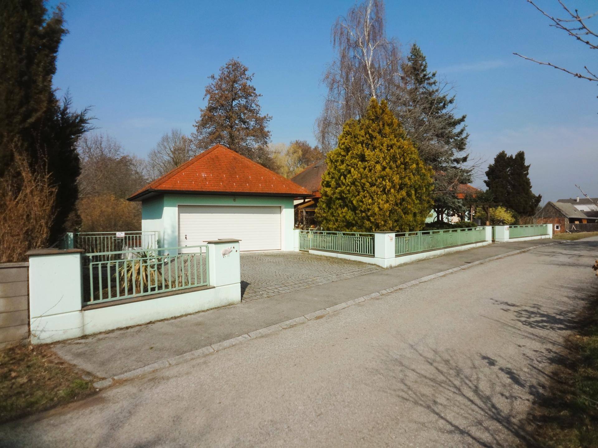 Garagenzufahrt mit grünem Zaun, gepflastertem Bereich und Bäumen.