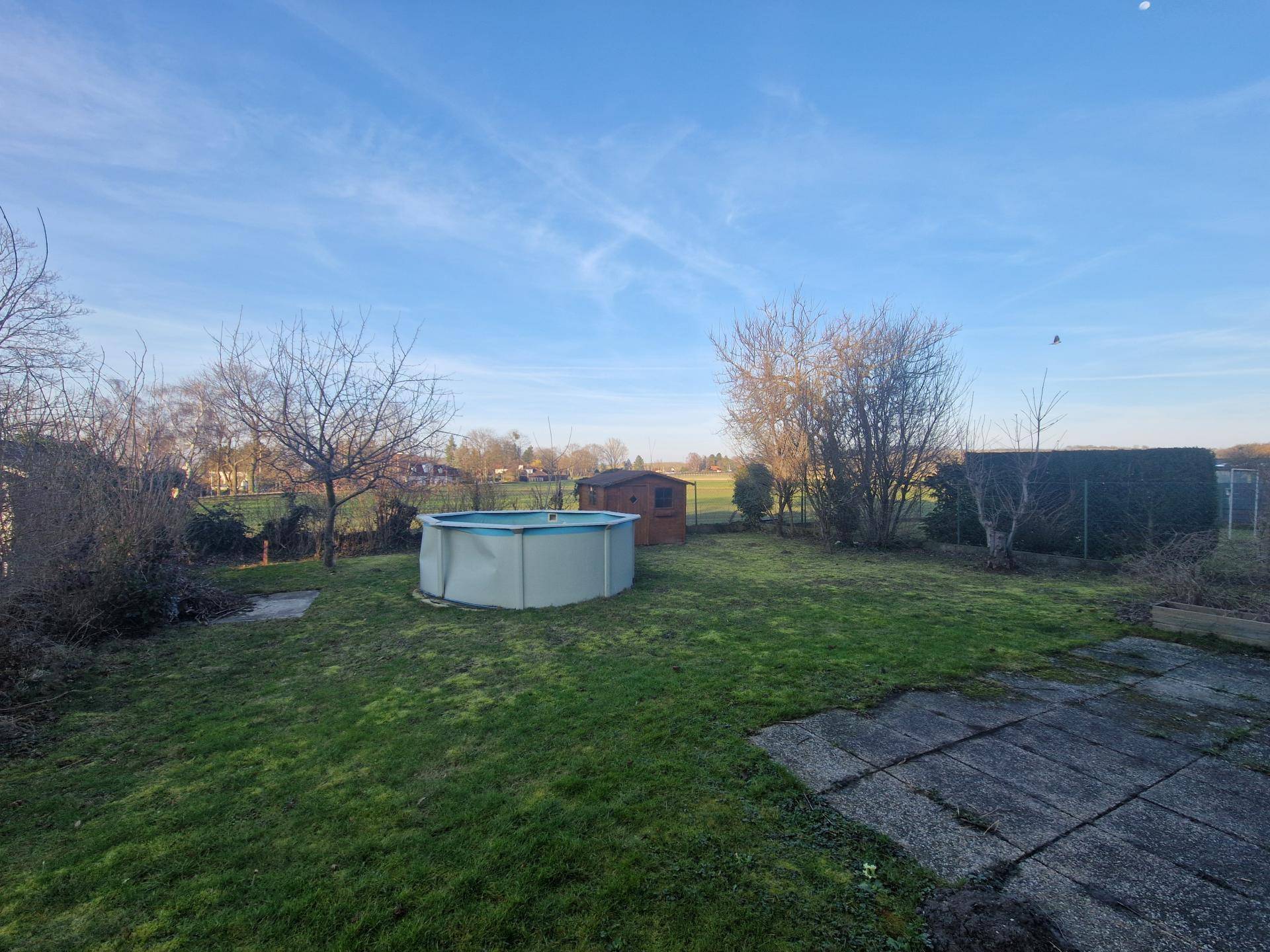 Großer Garten mit Rasenfläche, Aufstellpool und kleinem Gartenhaus unter blauem Himmel.
