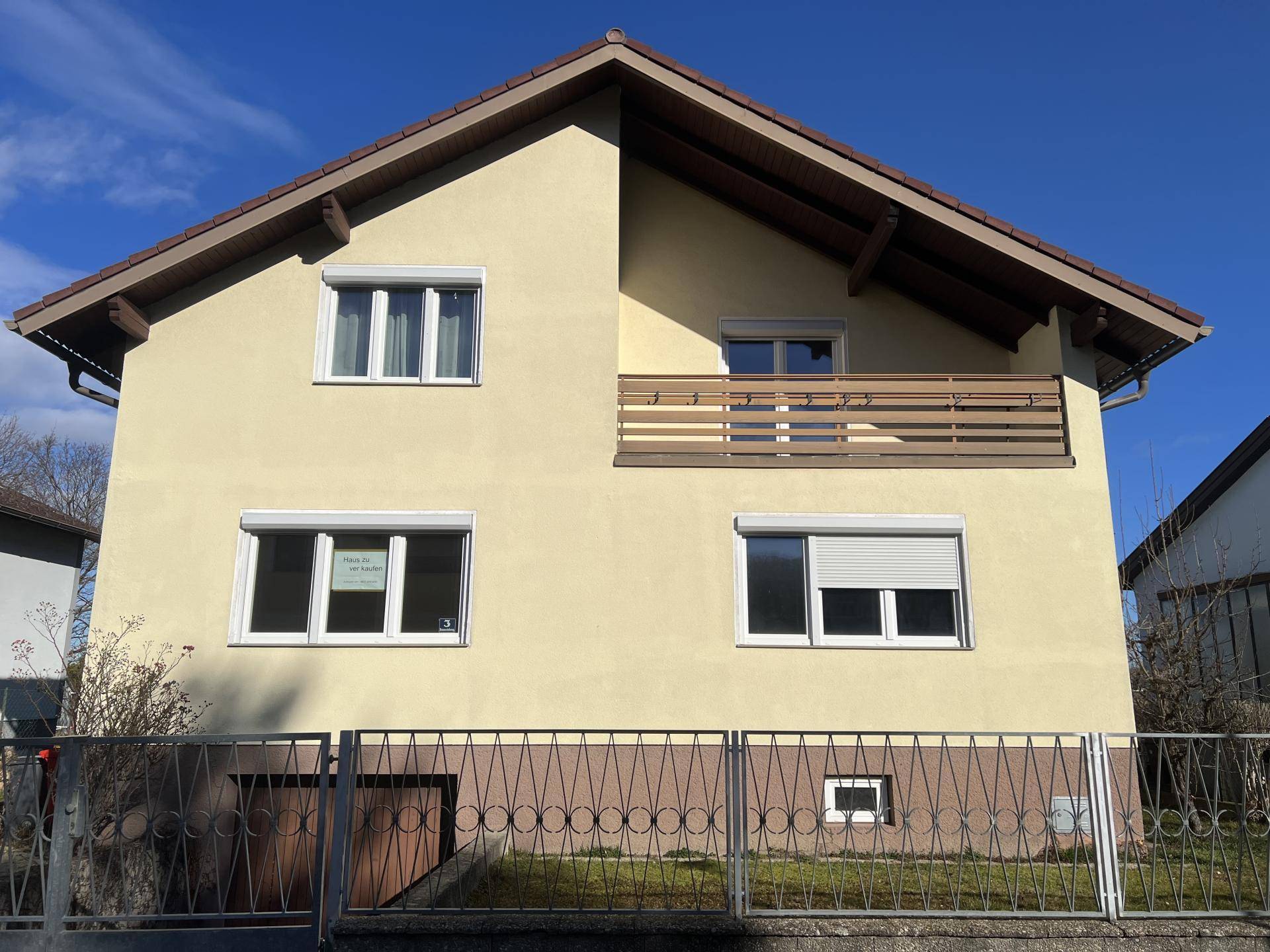 Vorderansicht des gelben Hauses mit Balkon und geschlossenen Rollläden an einem sonnigen Tag.