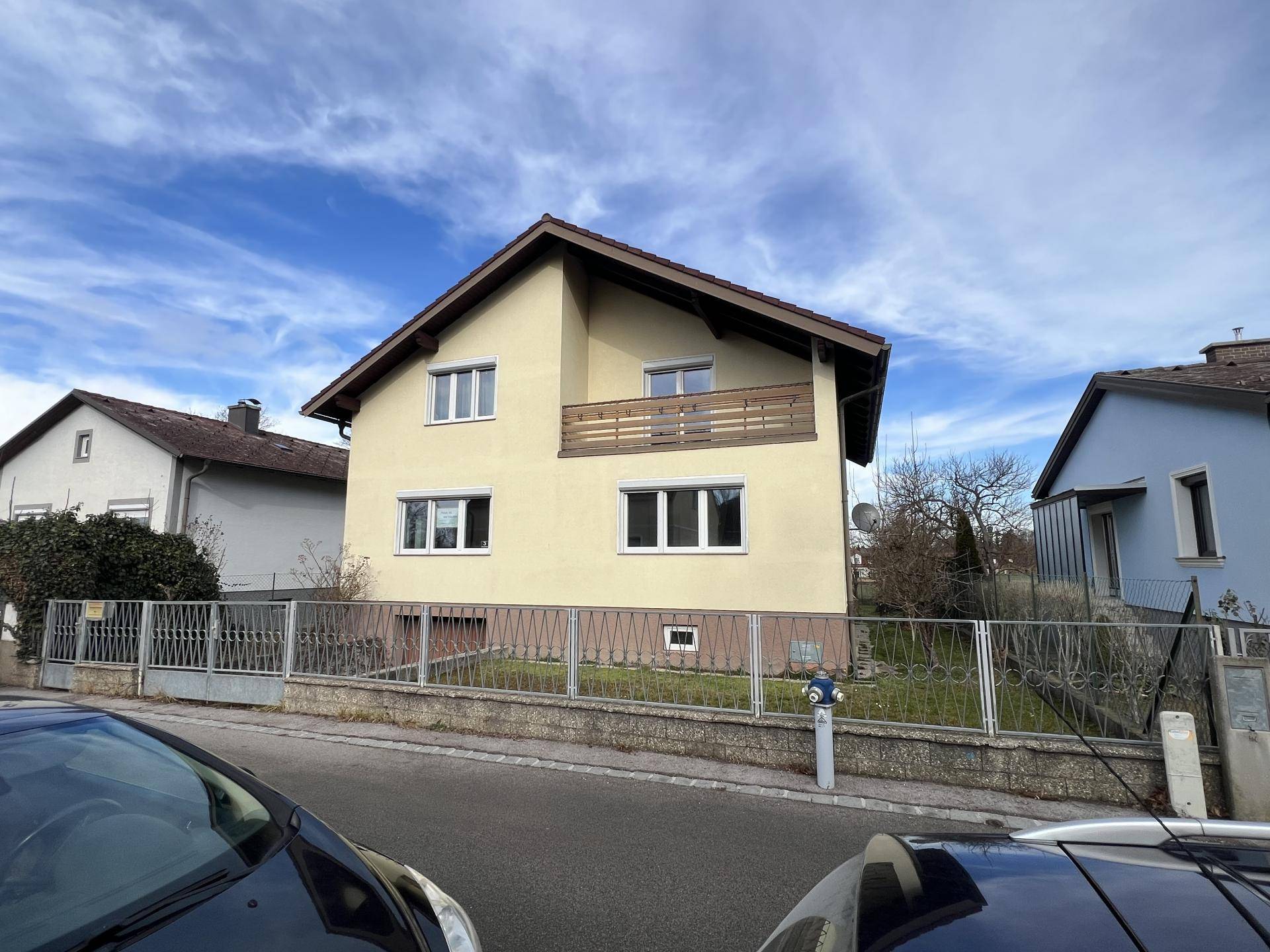 Einfamilienhaus von der Straße aus gesehen mit Vorgarten und Zaun, unter blauem Himmel.
