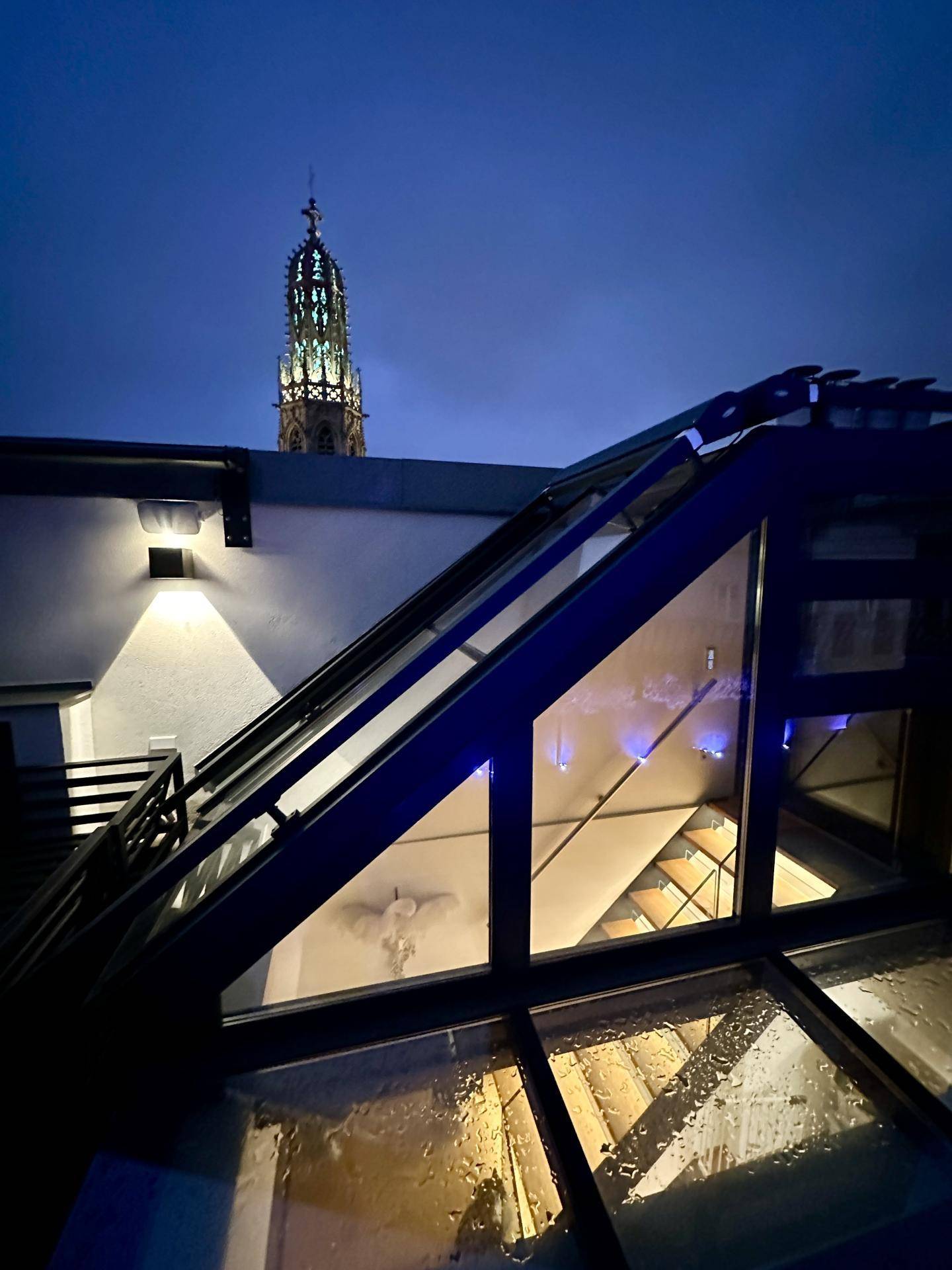 Nächtlicher Blick von der Dachterrasse auf einen beleuchteten Kirchturm unter dunklem Himmel.